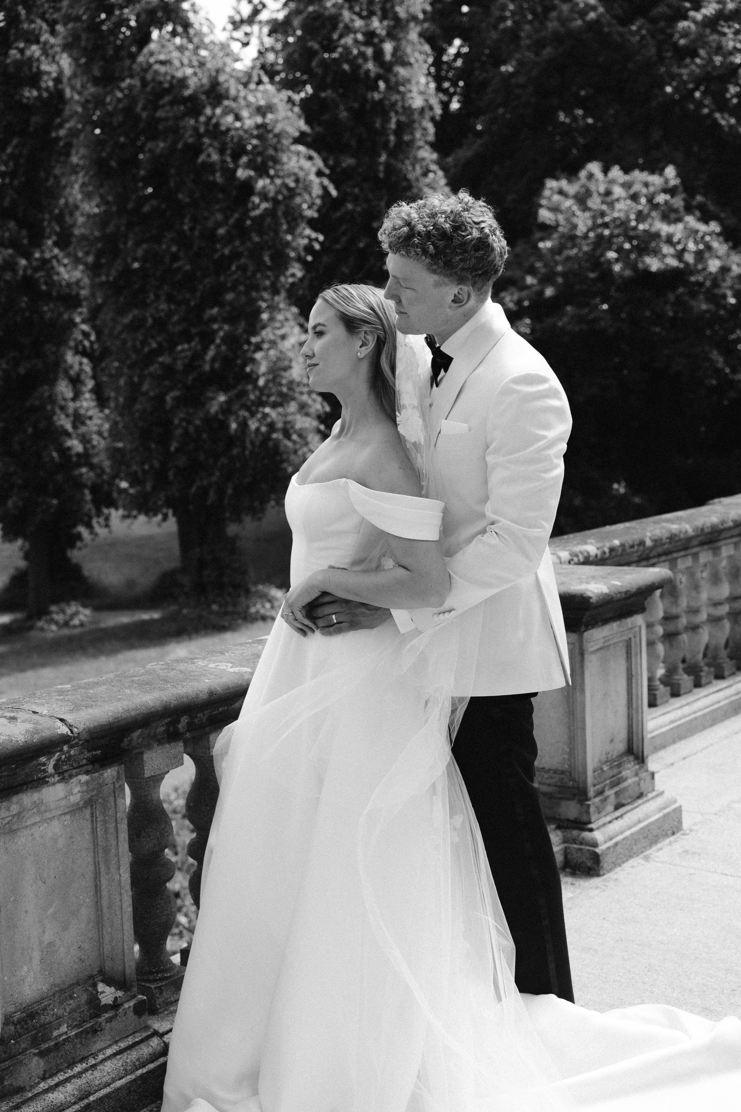 Black and white photograph of a bride and groom on their wedding day standing on a stone balcony outdoors with trees in the background. The bride is in a strapless wedding gown with puffed sleeves and a veil, while the groom is in a white tuxedo with