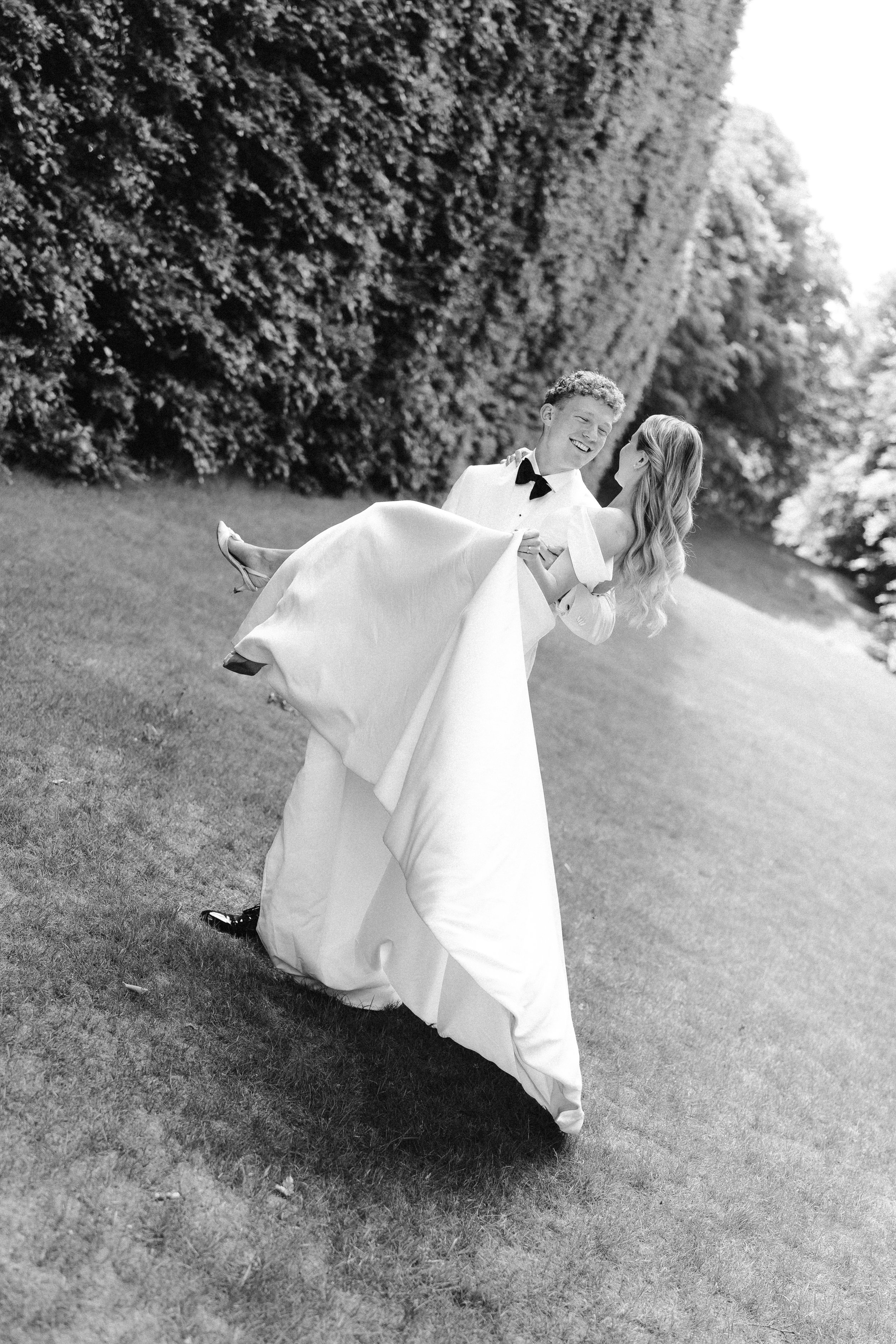 Black and white photo of a bride and groom outdoors, with the groom lifting the bride, both smiling, surrounded by trees.