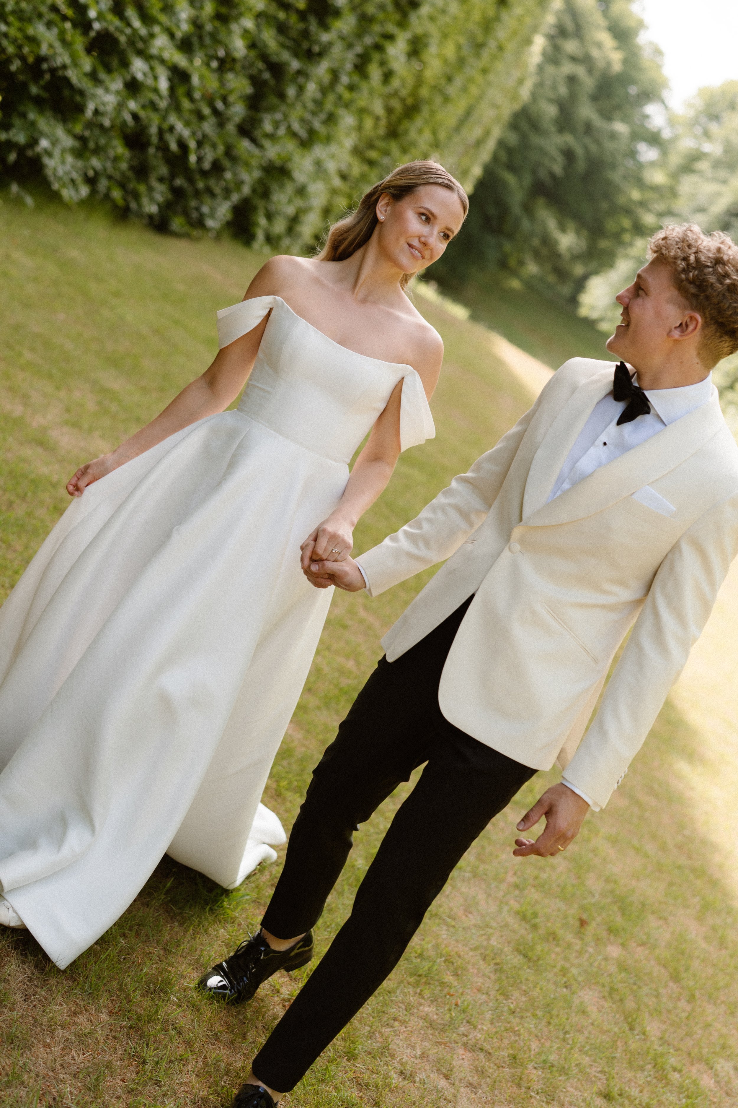 A bride and groom walking hand in hand outdoors, with the bride in a white wedding gown and the groom in a white tuxedo jacket with black pants and a bow tie.
