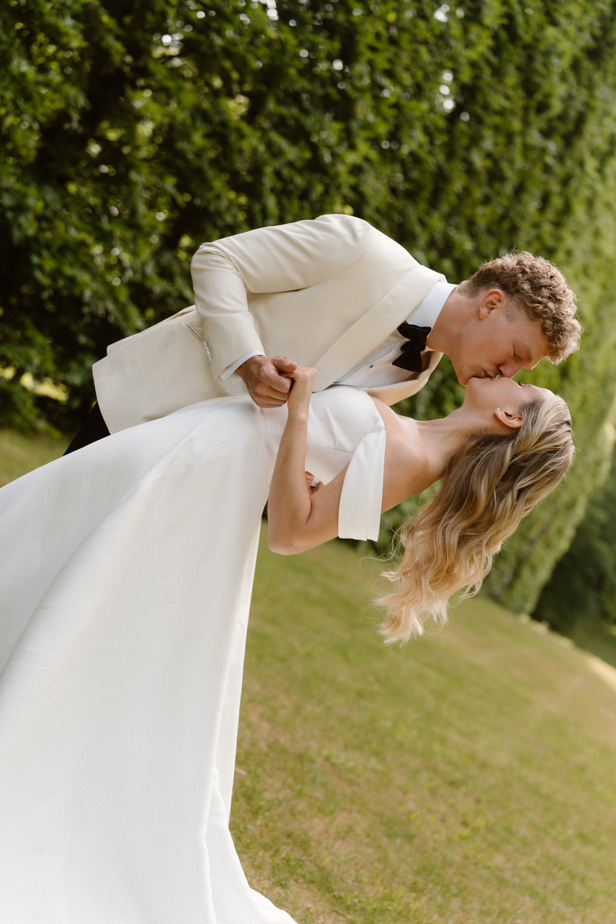 A bride and groom sharing a romantic kiss outdoors on their wedding day, holding hands, with green bushes in the background.