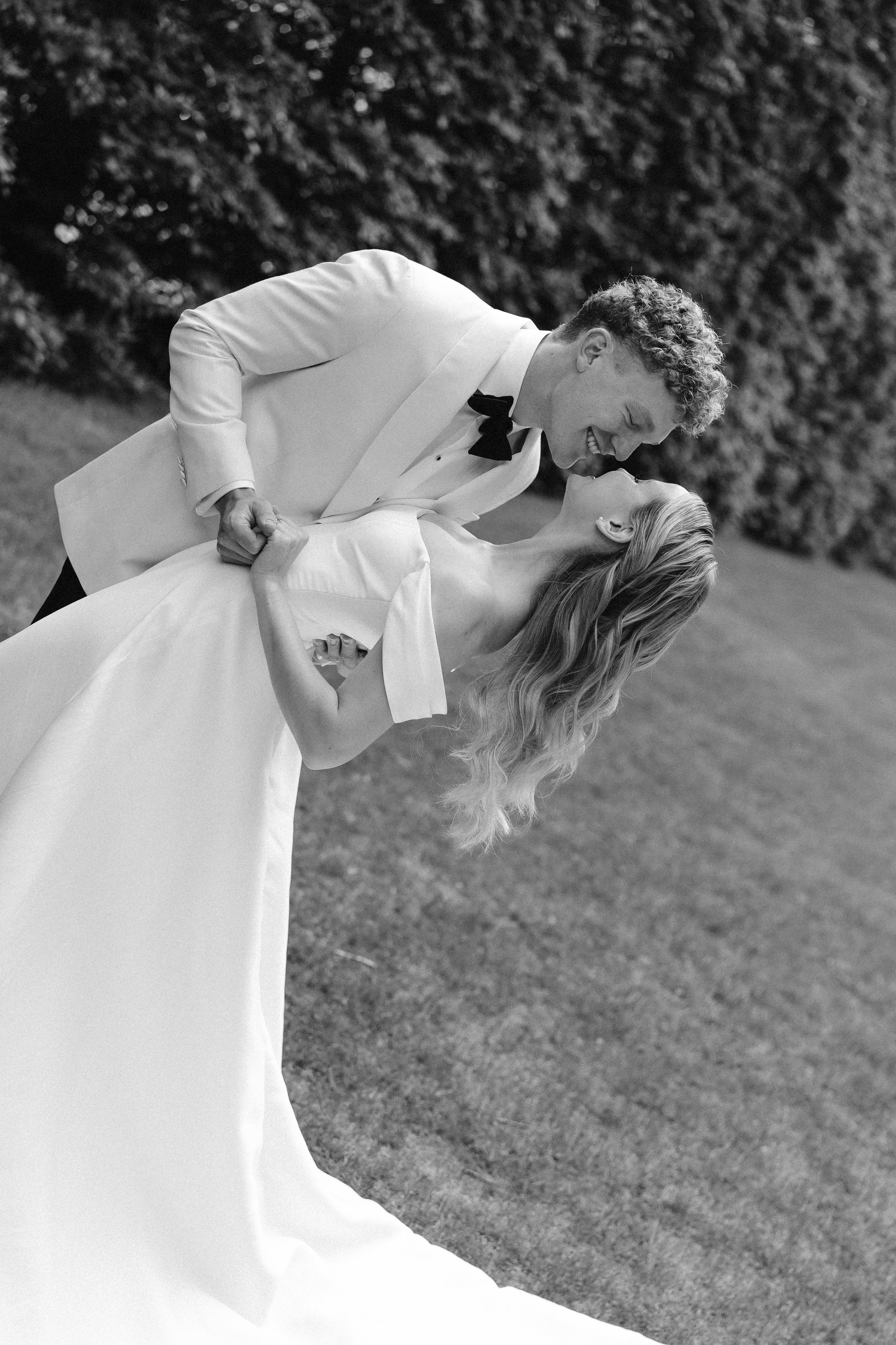 Black and white photo of a couple in wedding attire, possibly dancing, outdoors, with the man leaning over and smiling at the woman, who is holding his hand and tilting her head back.