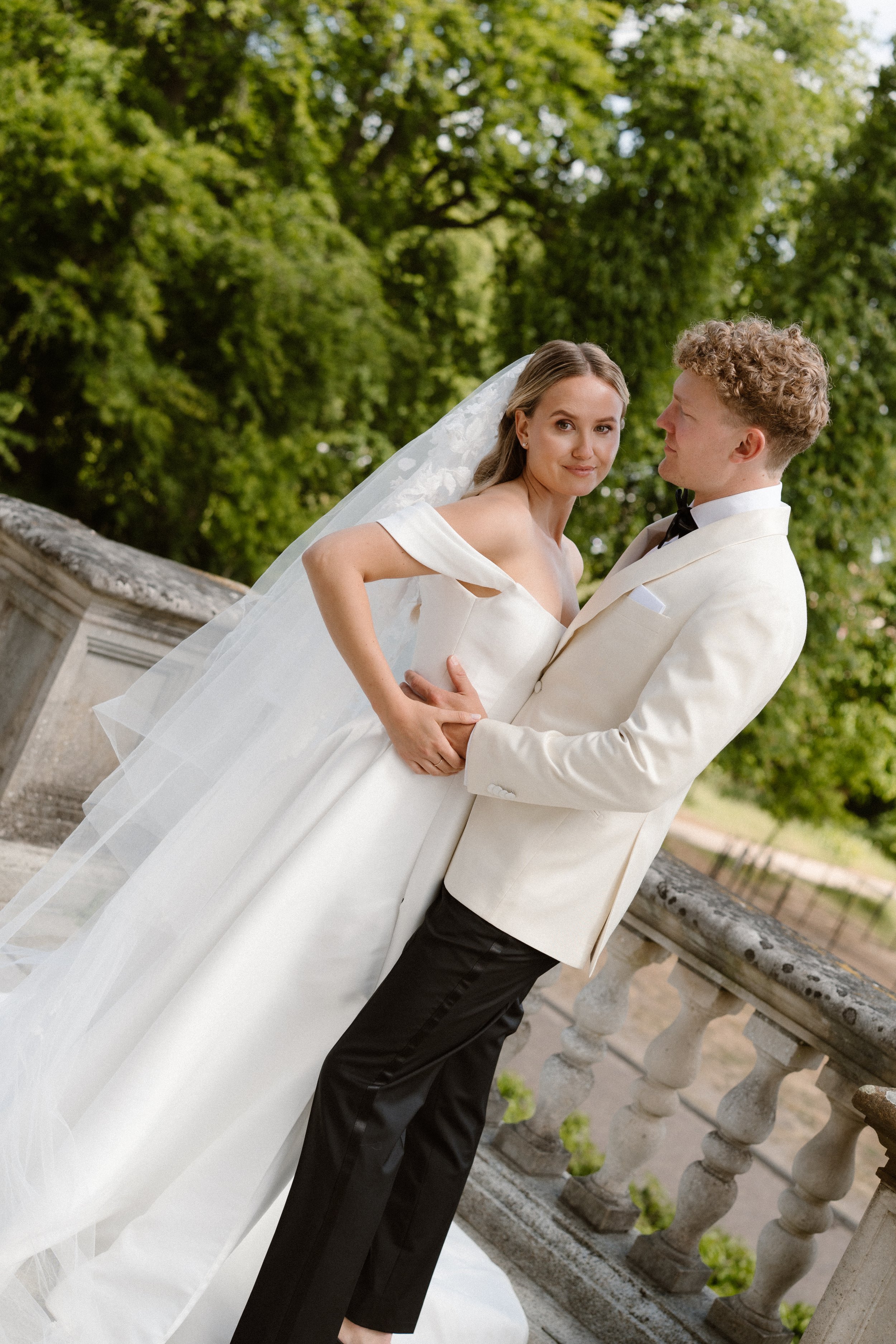 A bride and groom in wedding attire standing on a stone balcony outdoors with greenery in the background.