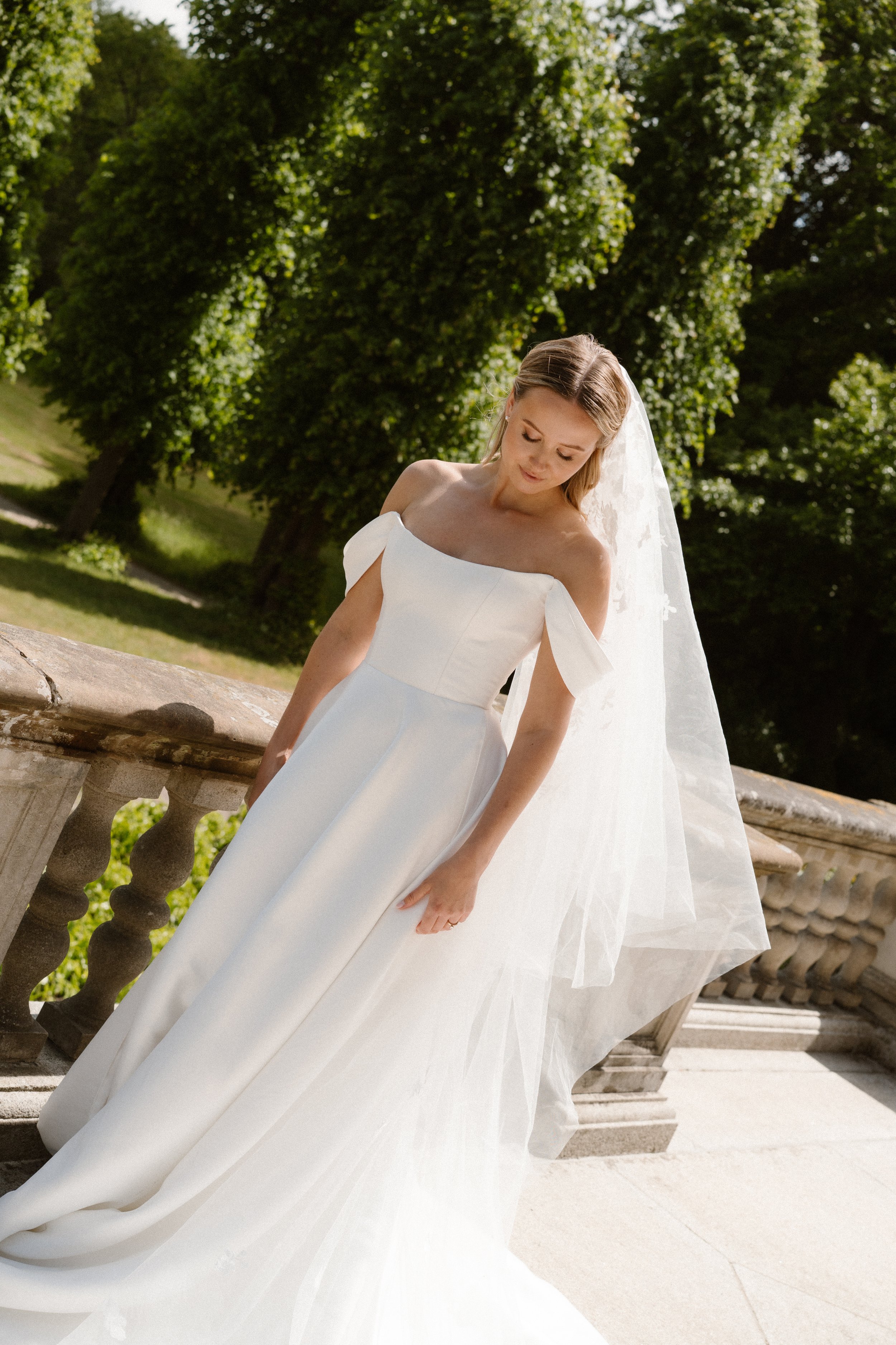 Bride in a white wedding dress with off-the-shoulder sleeves, standing on a stone balcony with green trees in the background.