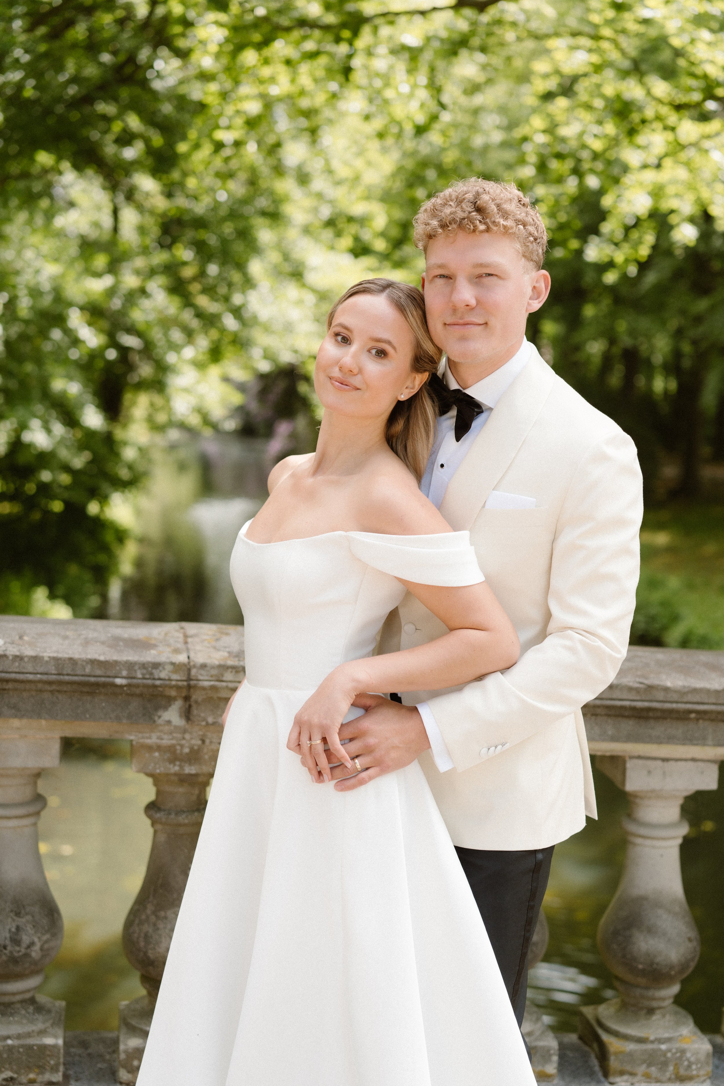 A bride and groom standing on a stone bridge in a park, with trees and a small waterfall in the background, during a wedding photo shoot.