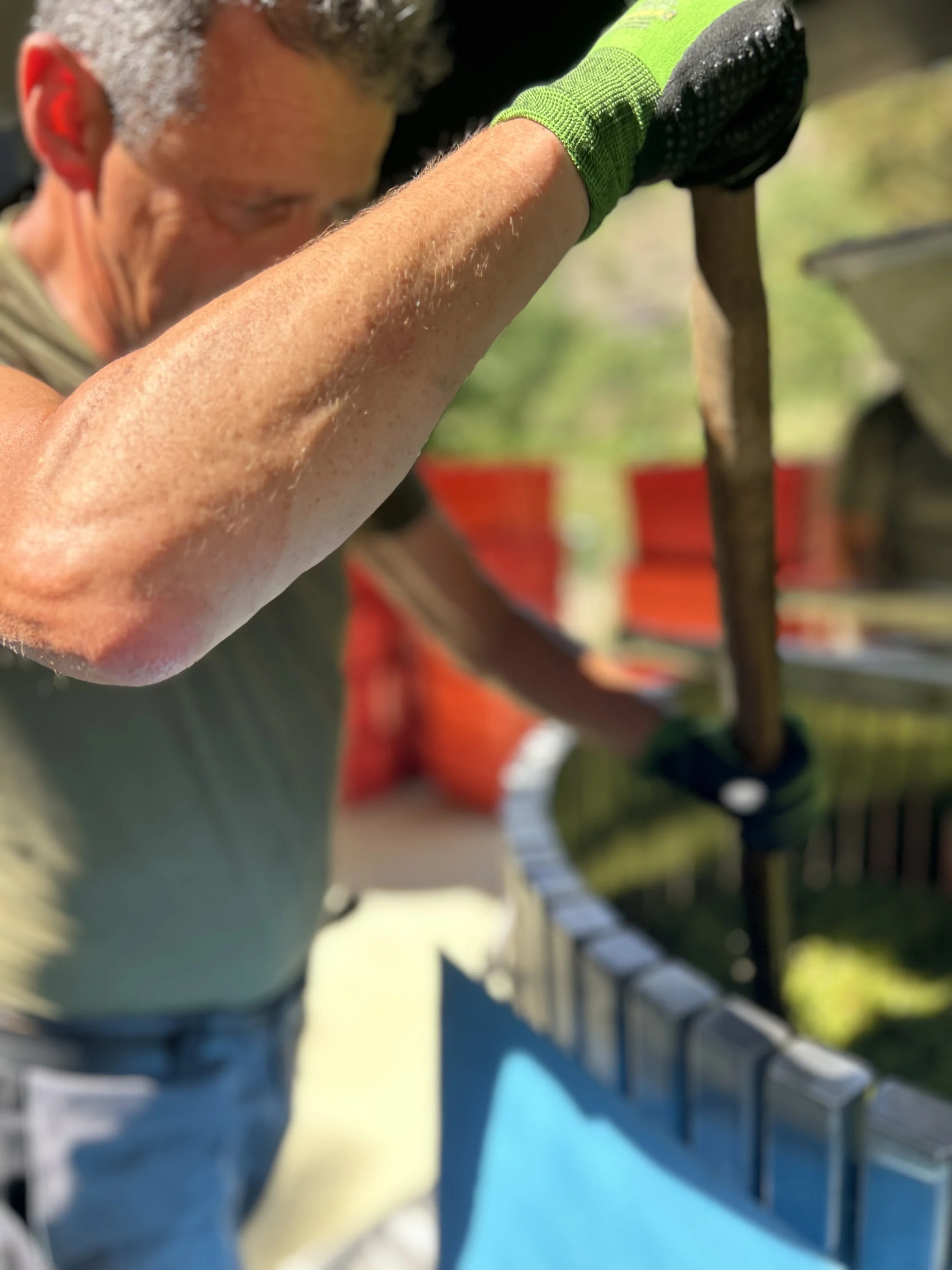 Ein Mann mit grauem Haar trägt einen grünen Handschuh und benutzt ein Werkzeug, wahrscheinlich einen Schraubenzieher, um an einer Batterie zu arbeiten. Im Hintergrund sind Orange- und Grünflächen sichtbar.