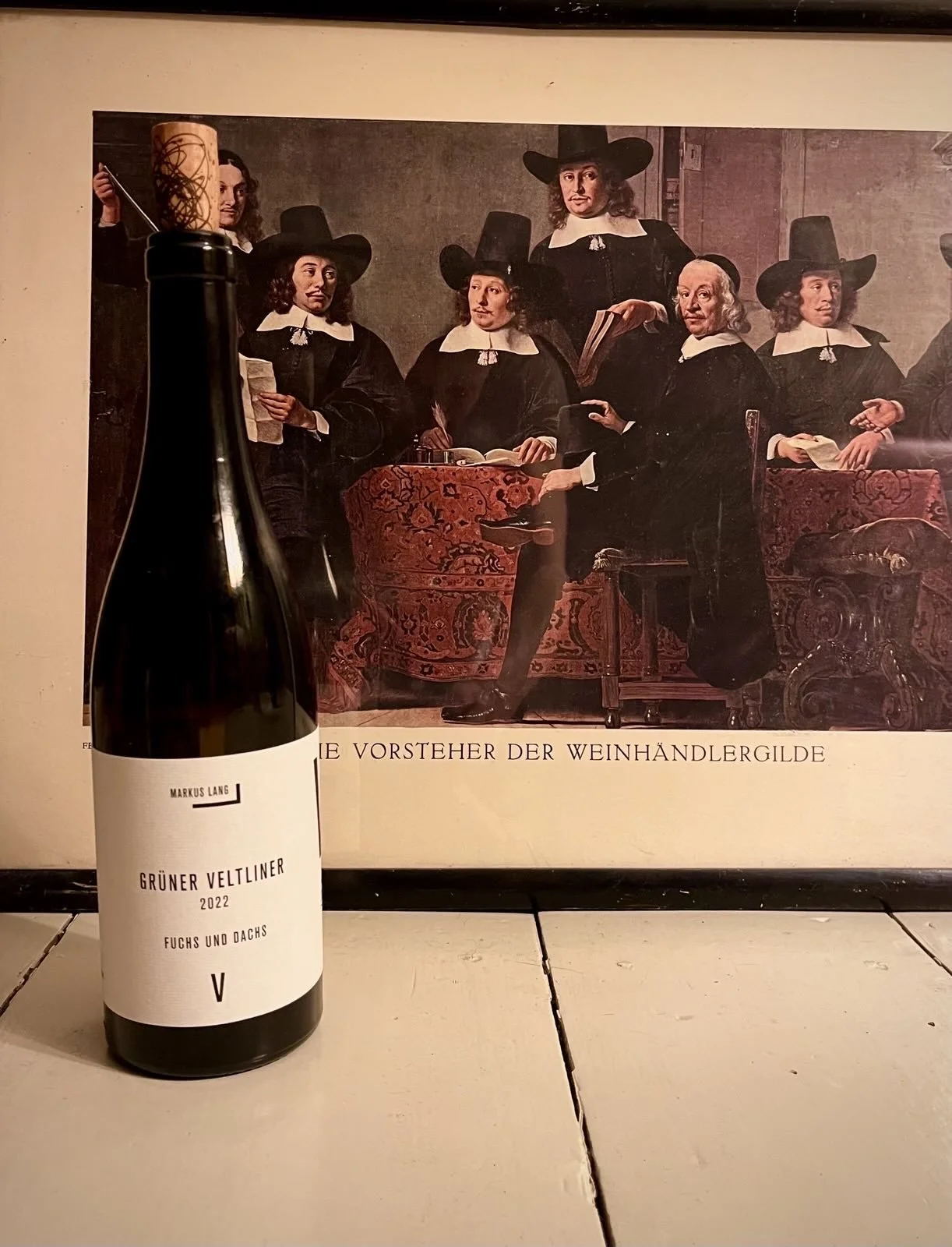Ein Weinflasche neben einem Kunstdruck mit einer Gruppe von Männern im Barockstil, die in schwarzen Kleidern und Hüten abgebildet sind.
