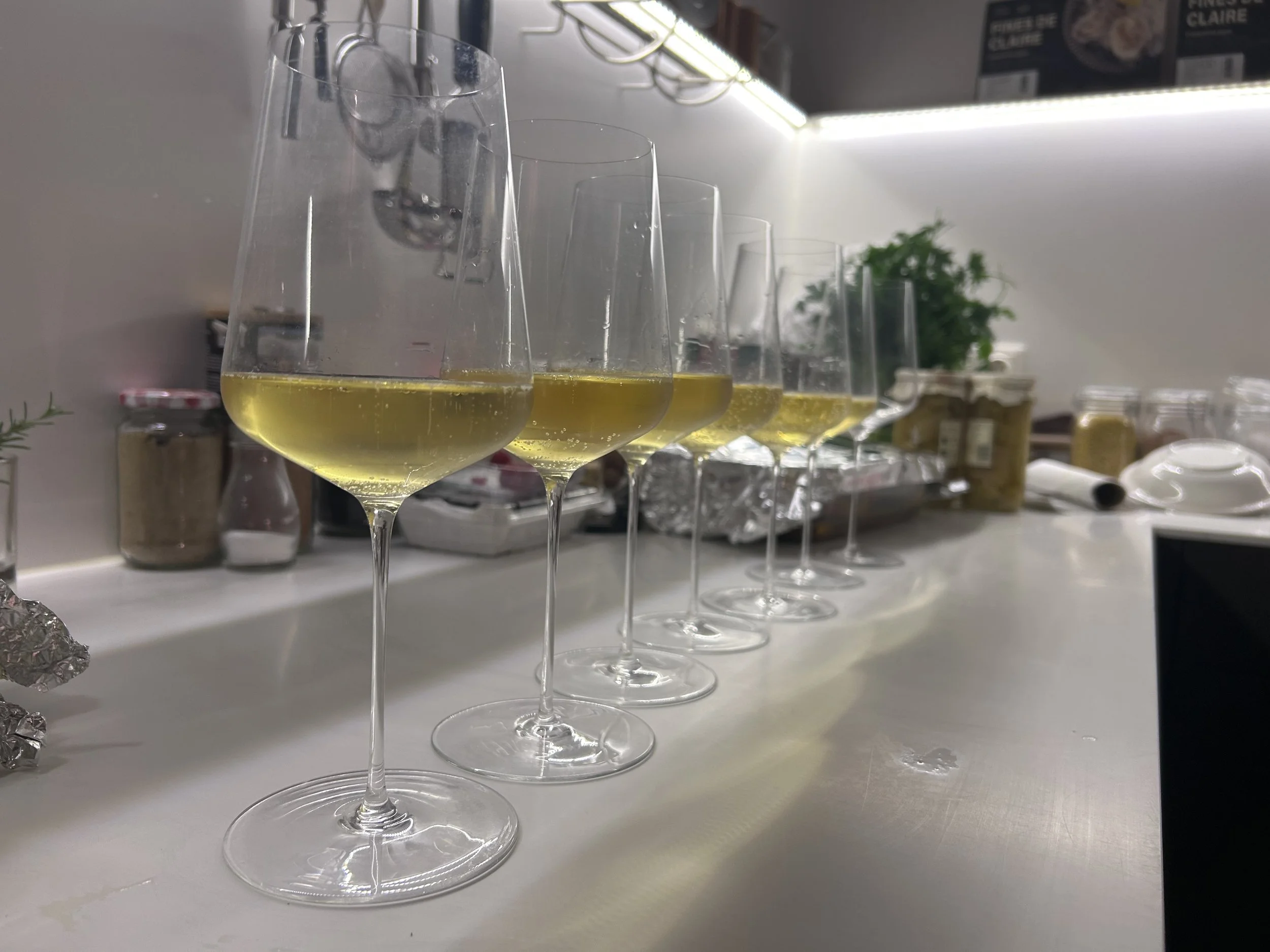 Mehrere Weingläser mit weißem Wein auf einer weißen Theke im Hintergrund sehen man Küchenutensilien, Gläser, Dosen und einen Topf mit einer Pflanze.