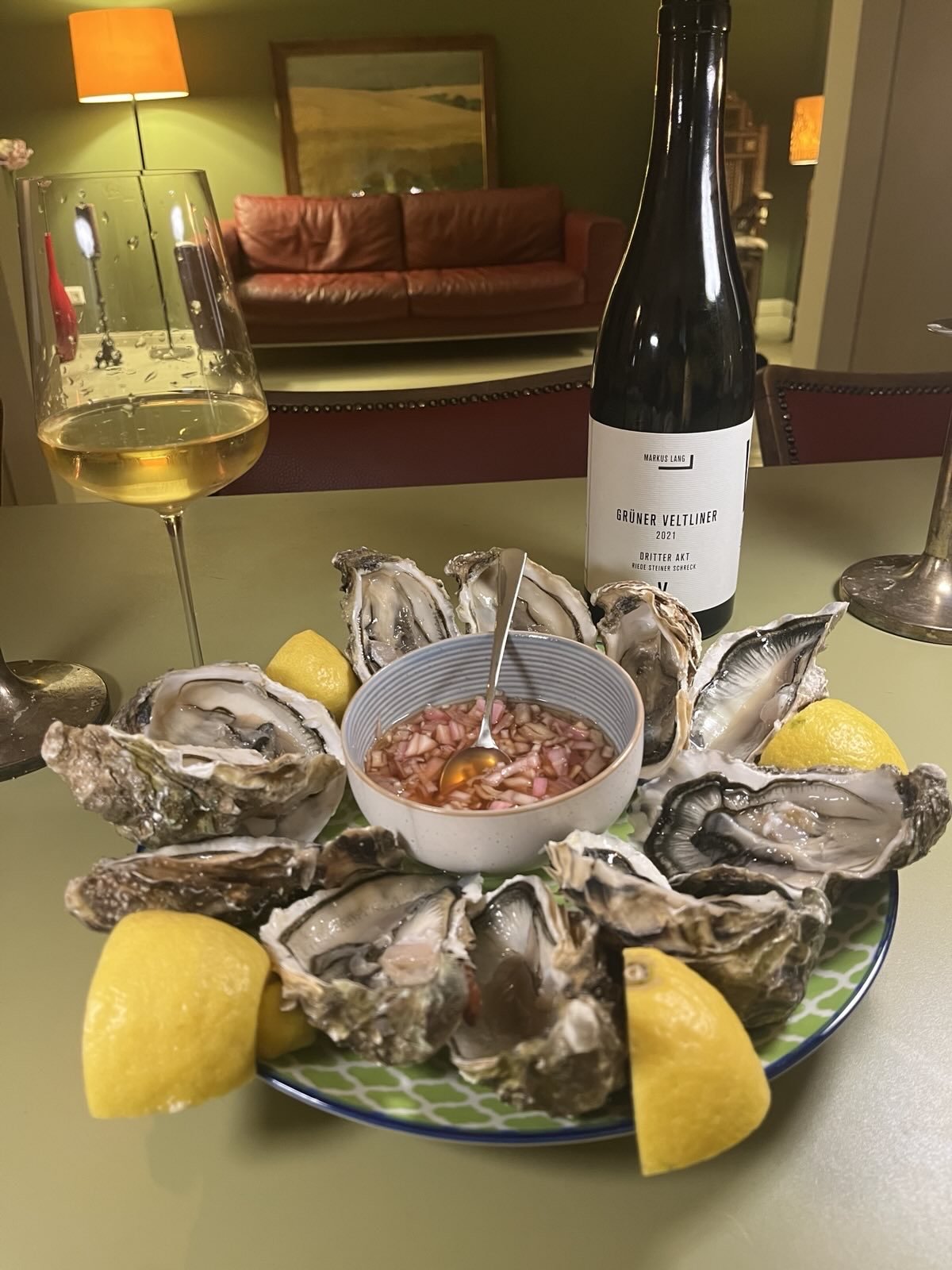 Ein Teller mit frischen Austern, Zitronenspalten und einer Schüssel Austernsoße auf einem Tisch, im Hintergrund ein Glas Weißwein, eine Flasche Wein, ein Wohnzimmer mit grünen Wänden, einem roten Sofa, einer stehenden Lampe und einem Gemälde an der W