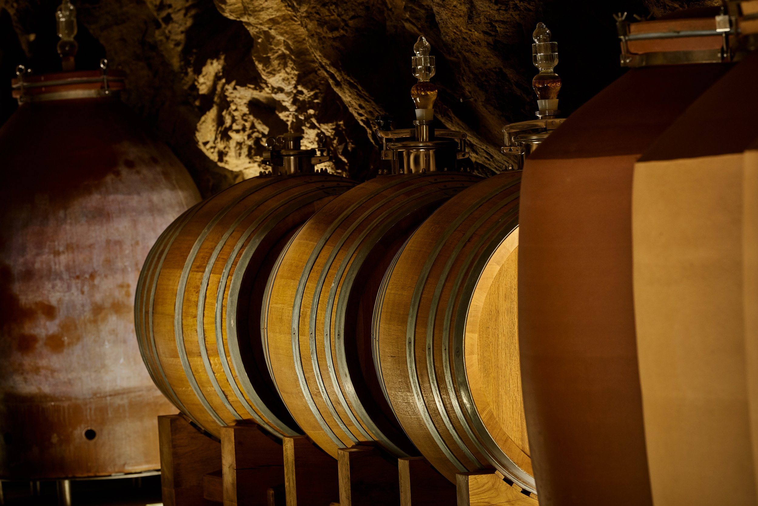 Holzfässer in einem Weinkeller, teilweise aus Metall mit Holzfellen und Edelstahlelementen, vor einer Felswand