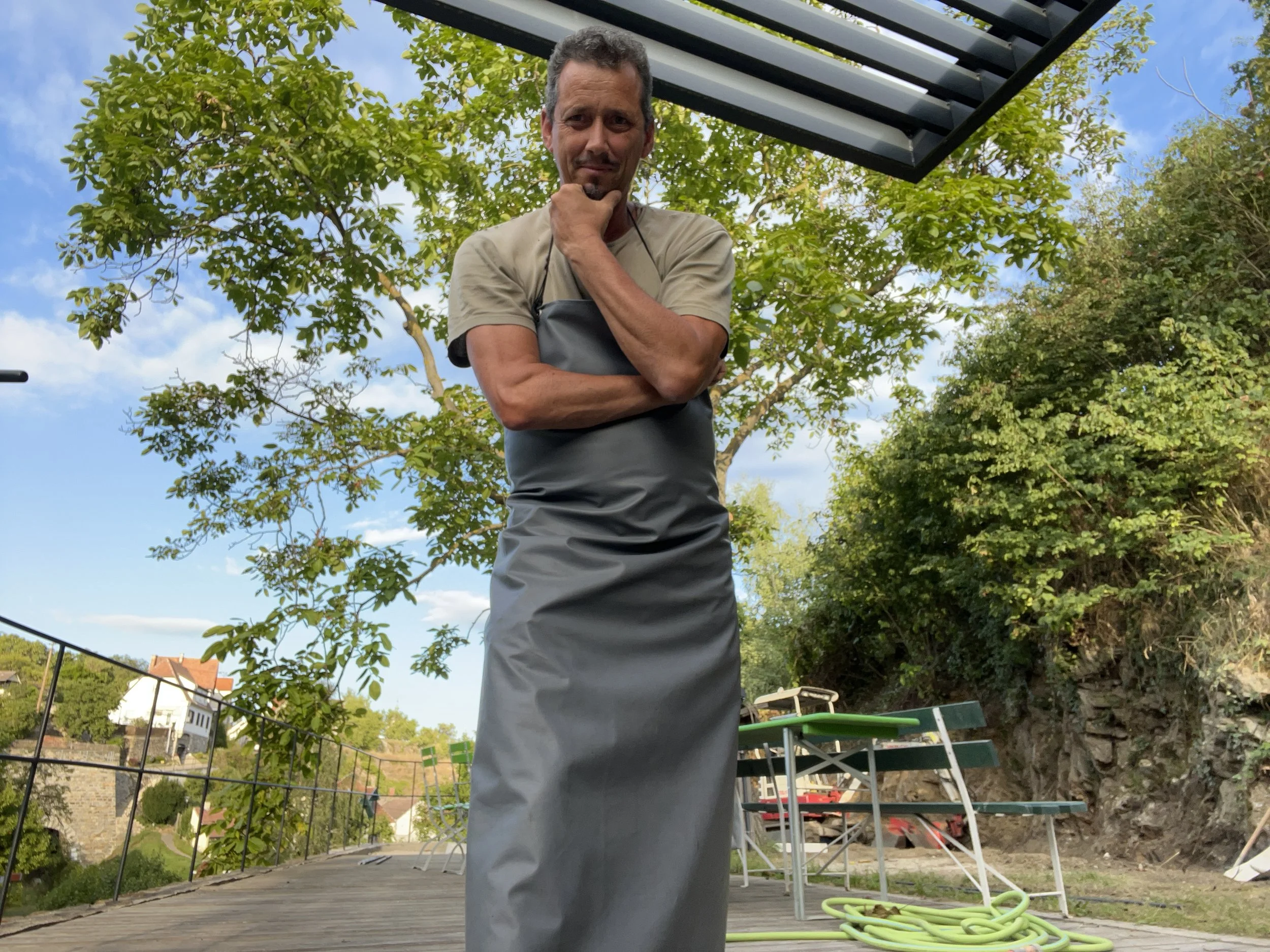 Ein Mann trägt eine graue Schürze und steht auf einer Terrasse im Freien, umgeben von Bäumen und Gartenmöbeln, mit einem blauen Himmel im Hintergrund.