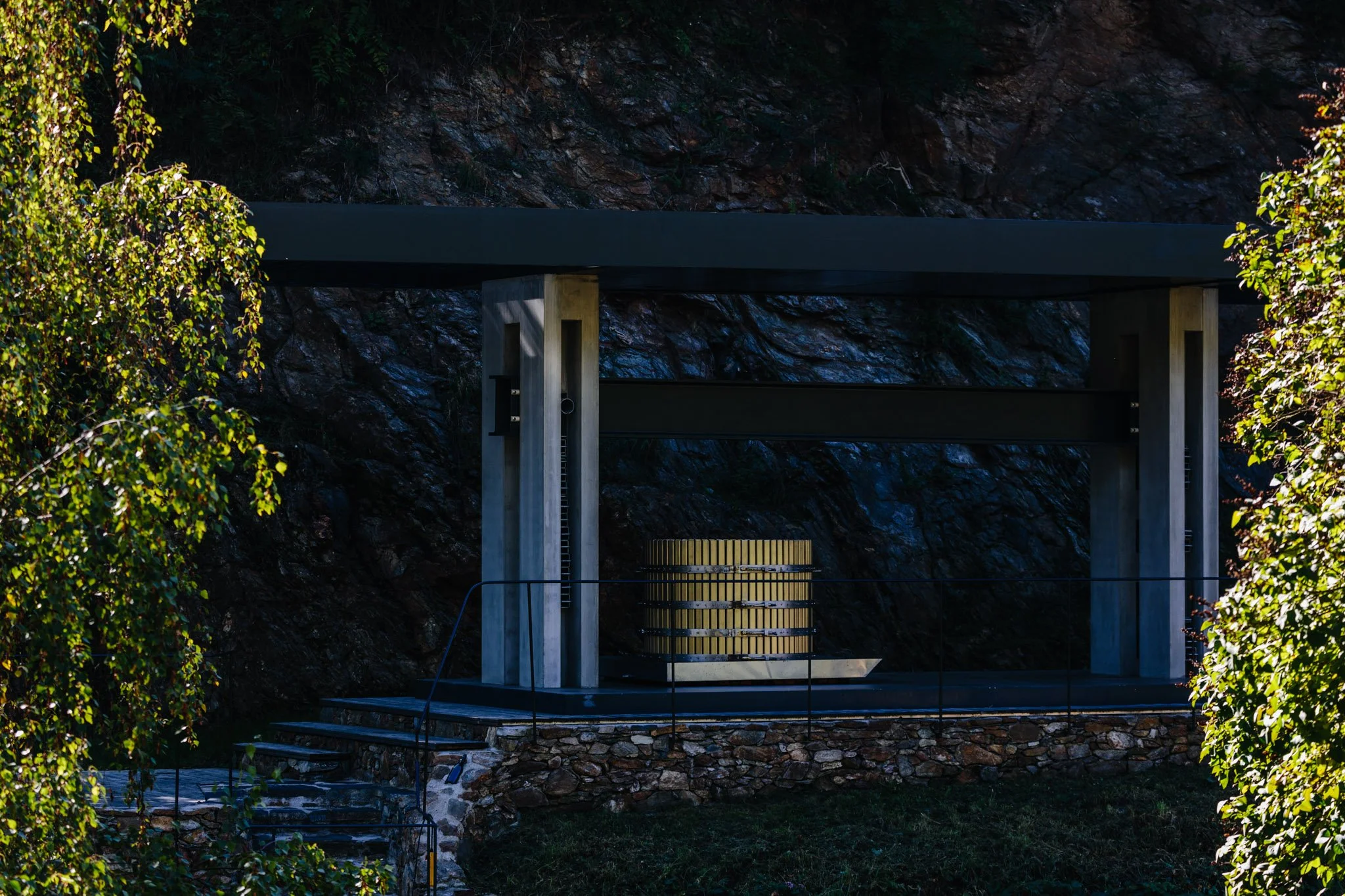 Moderner Waschturm aus Holz in einer kleinen, offenen, überdachten Struktur mit Betonstützen, eingebettet in die Natur zwischen Bäumen und Felsen.
