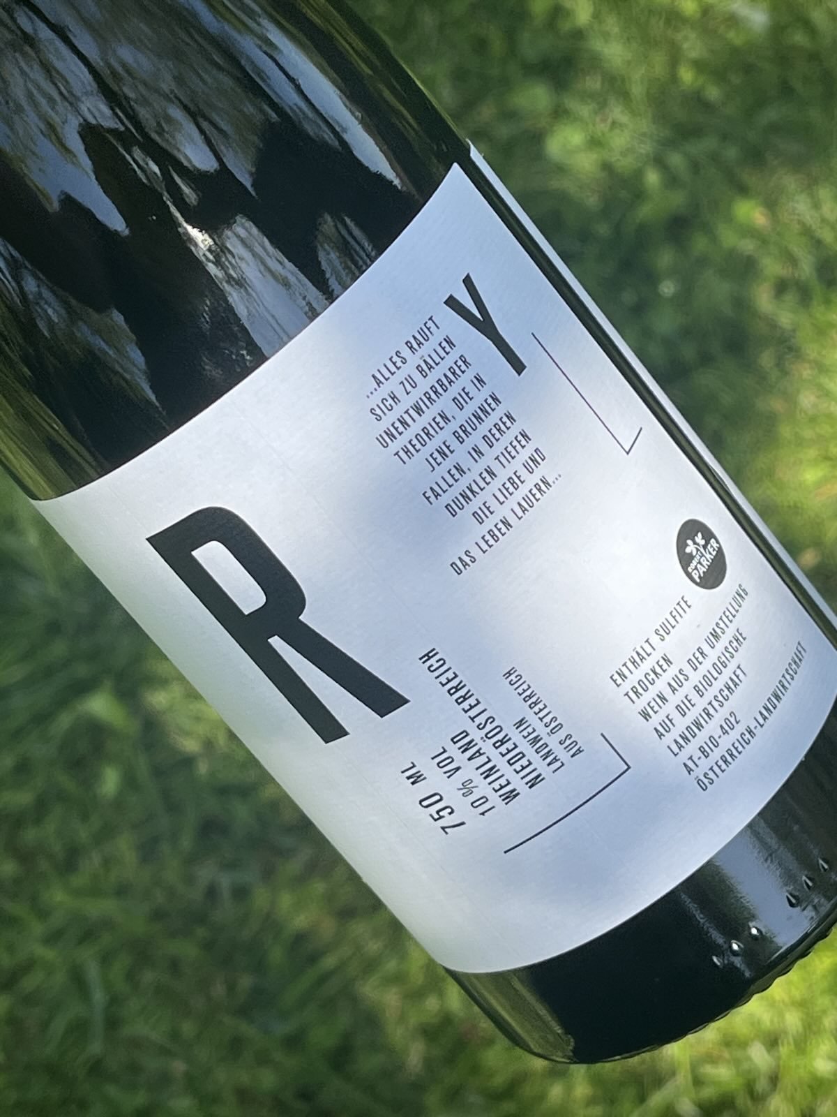 Schwarze Flasche mit weißem Etikett, das تحتويّ einen Text auf Deutsch und das Logo von Parfer. Die Flasche liegt auf grünem Gras.