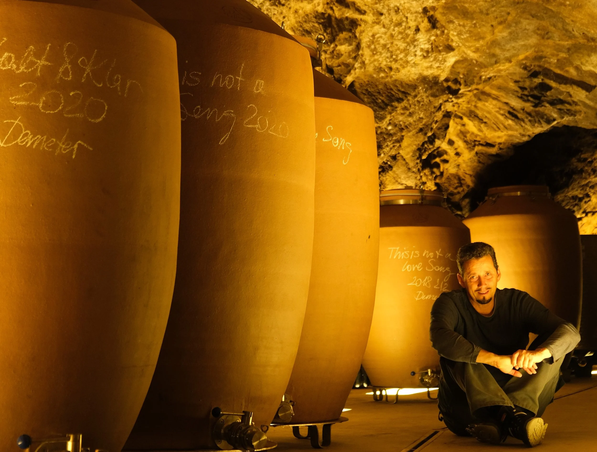 Ein Mann sitzt auf dem Boden in einem Keller neben großen, braunen Fässern. Die Wände sind aus Felsen. Das Licht ist warm und gelblich.