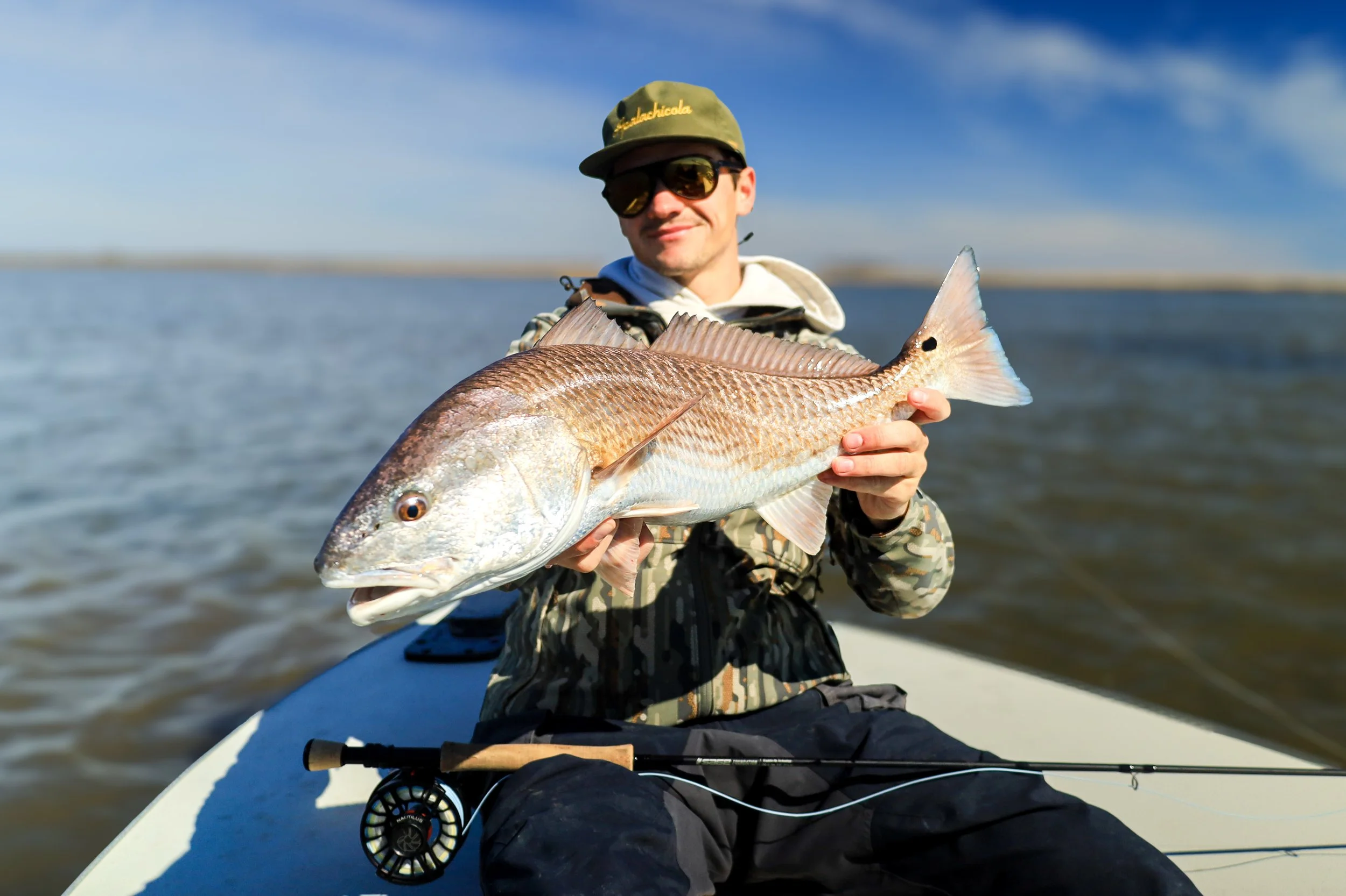 Man in camouflage jacket and baseball cap sitting in a boat, holding a large fish he caught, with fishing rod lying across his lap.