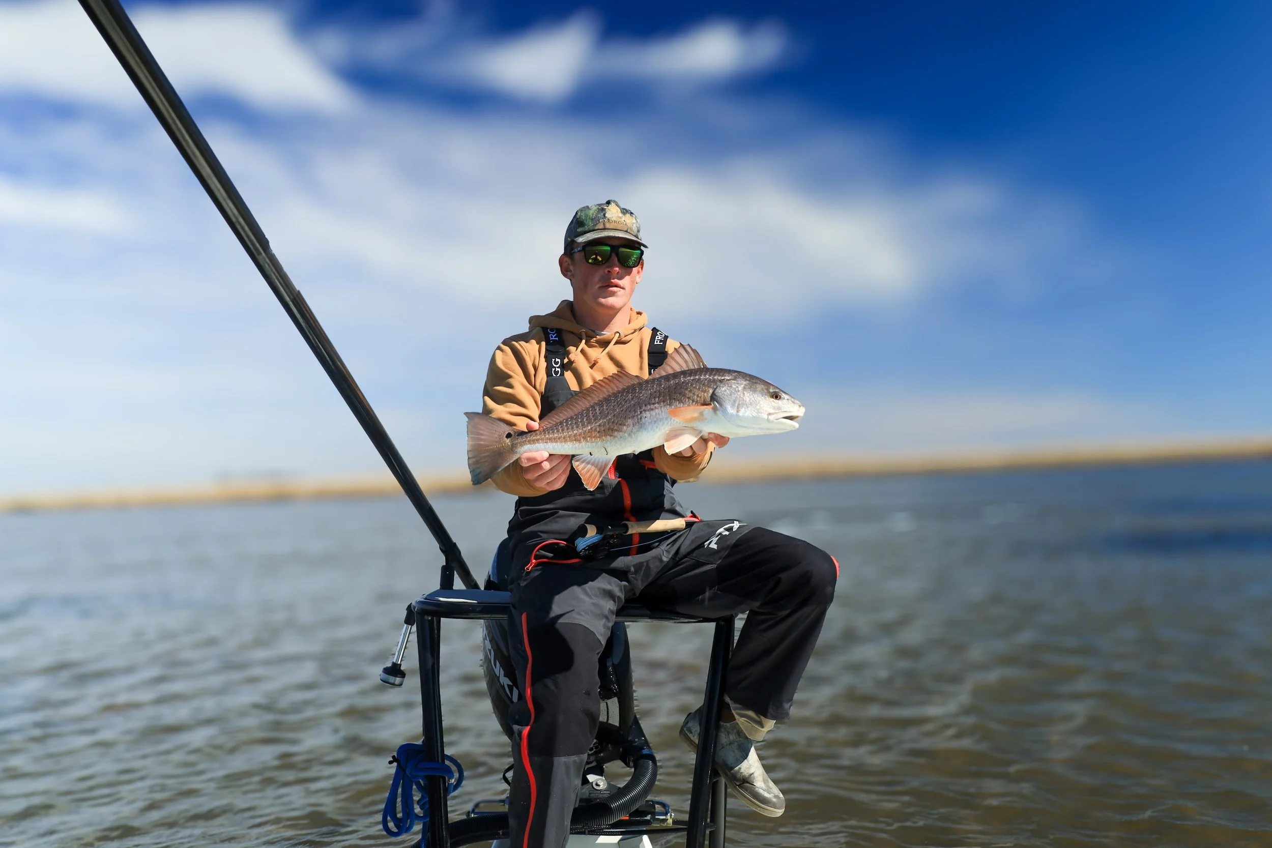 A young man sitting on a boat holding a large fish he caught, with water and a blue sky in the background.