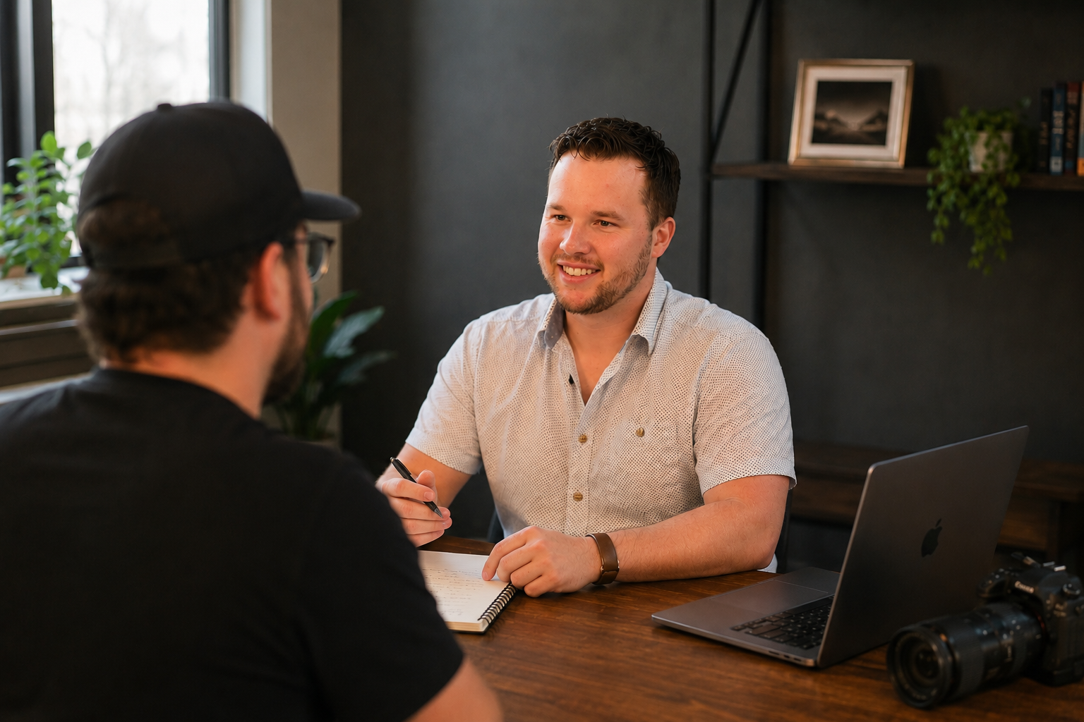 Two men having a conversation at a wooden desk in a modern office, one smiling, the other seen from behind wearing a black shirt and cap. On the desk are a laptop, a camera, and a notebook.