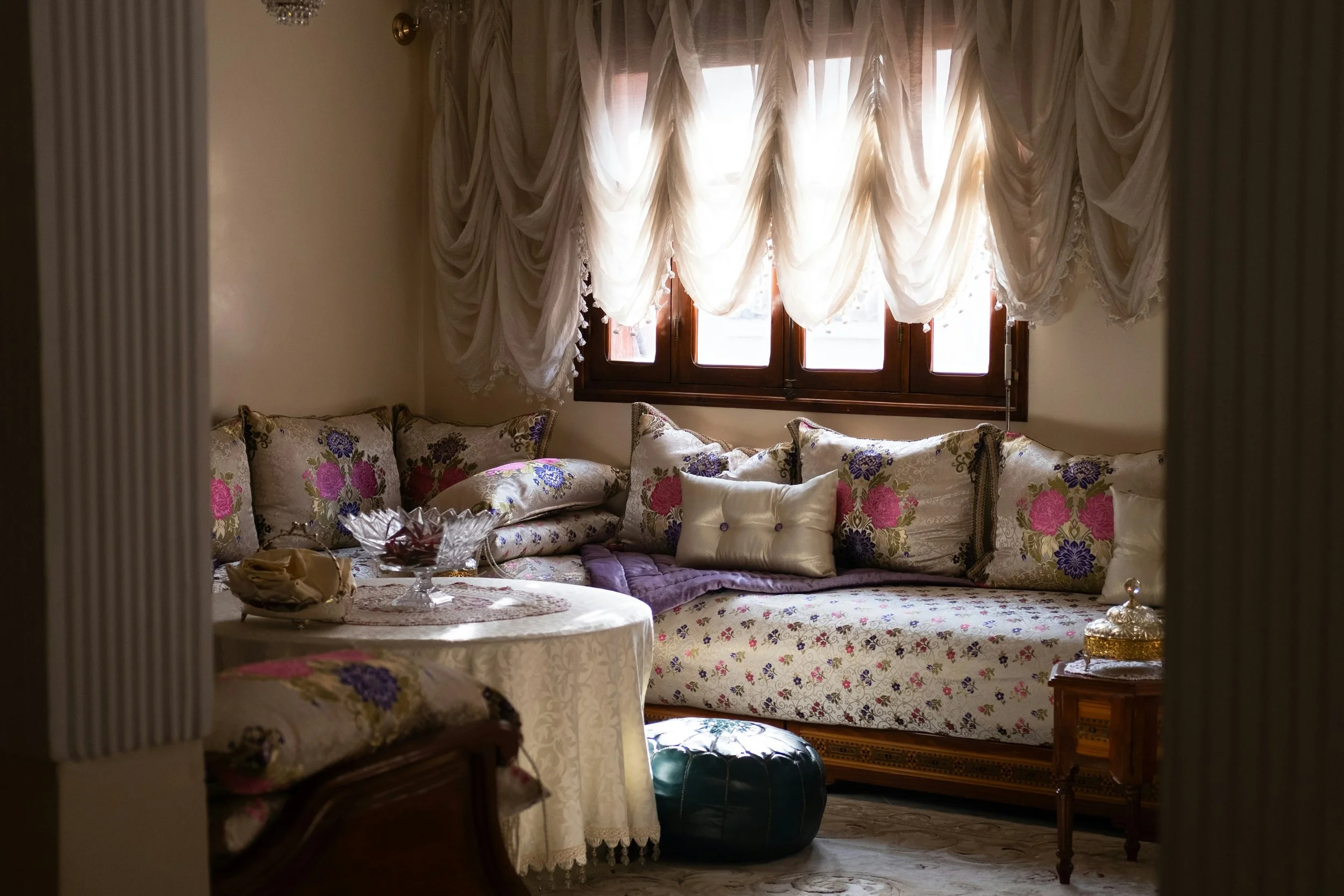 A cozy living room with a large floral-patterned sofa decorated with various pillows, a round table covered with a white tablecloth holding a glass bowl, and a window with elegant white curtains. There is a small wooden side table with a decorative jar and a black ottoman on the floor.