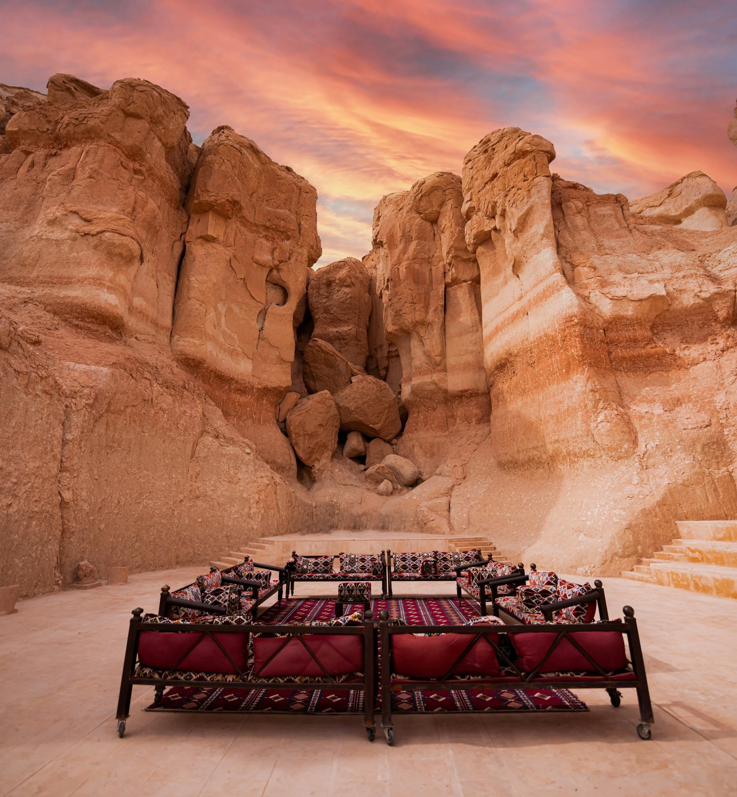 Arab floor seating area with patterned cushions and carpets surrounded by tall sandstone cliffs during sunset.