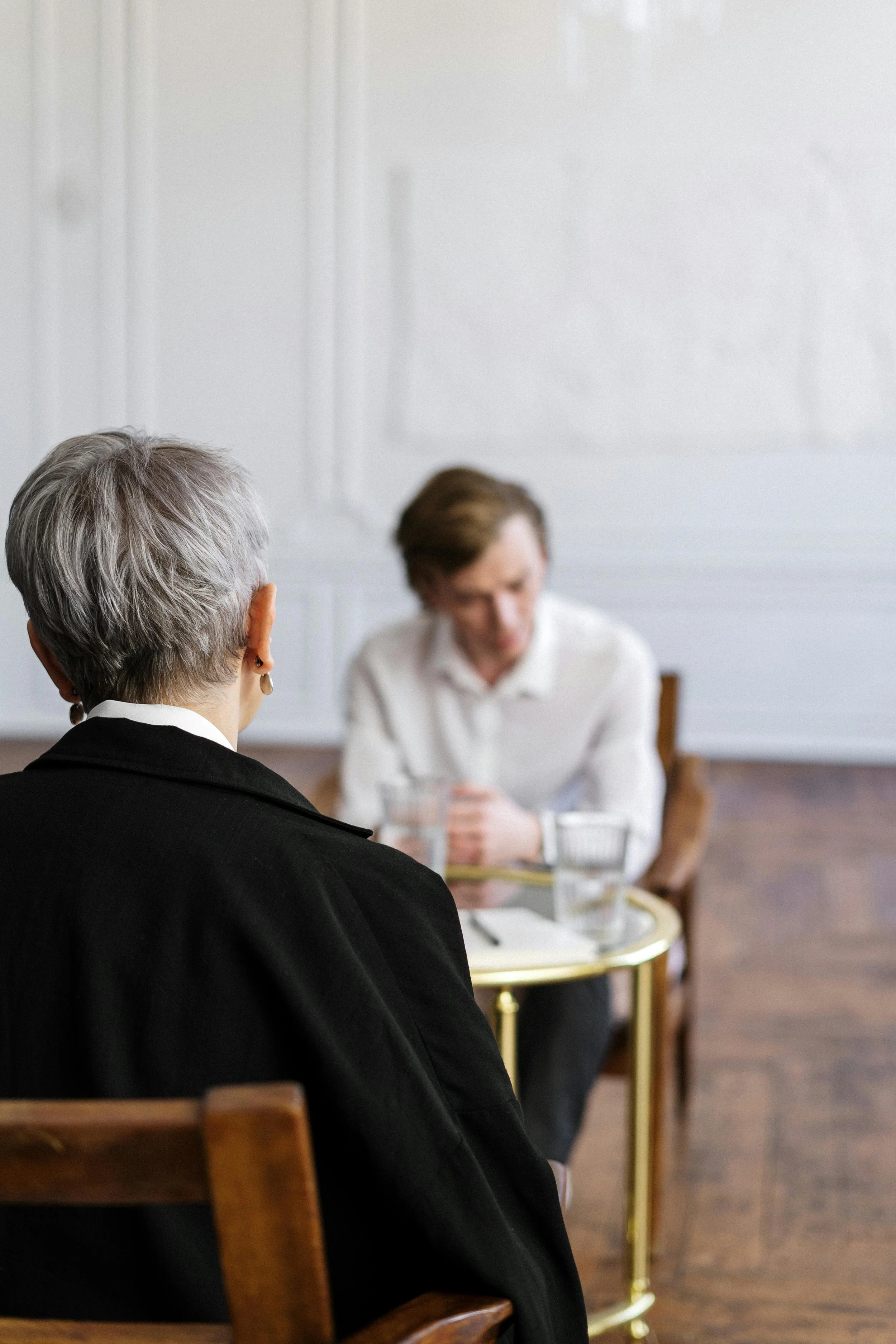 An older woman with gray hair and earrings sits facing a younger man with brown hair, at a small round table with glasses of water, during a serious conversation in a well-lit room.