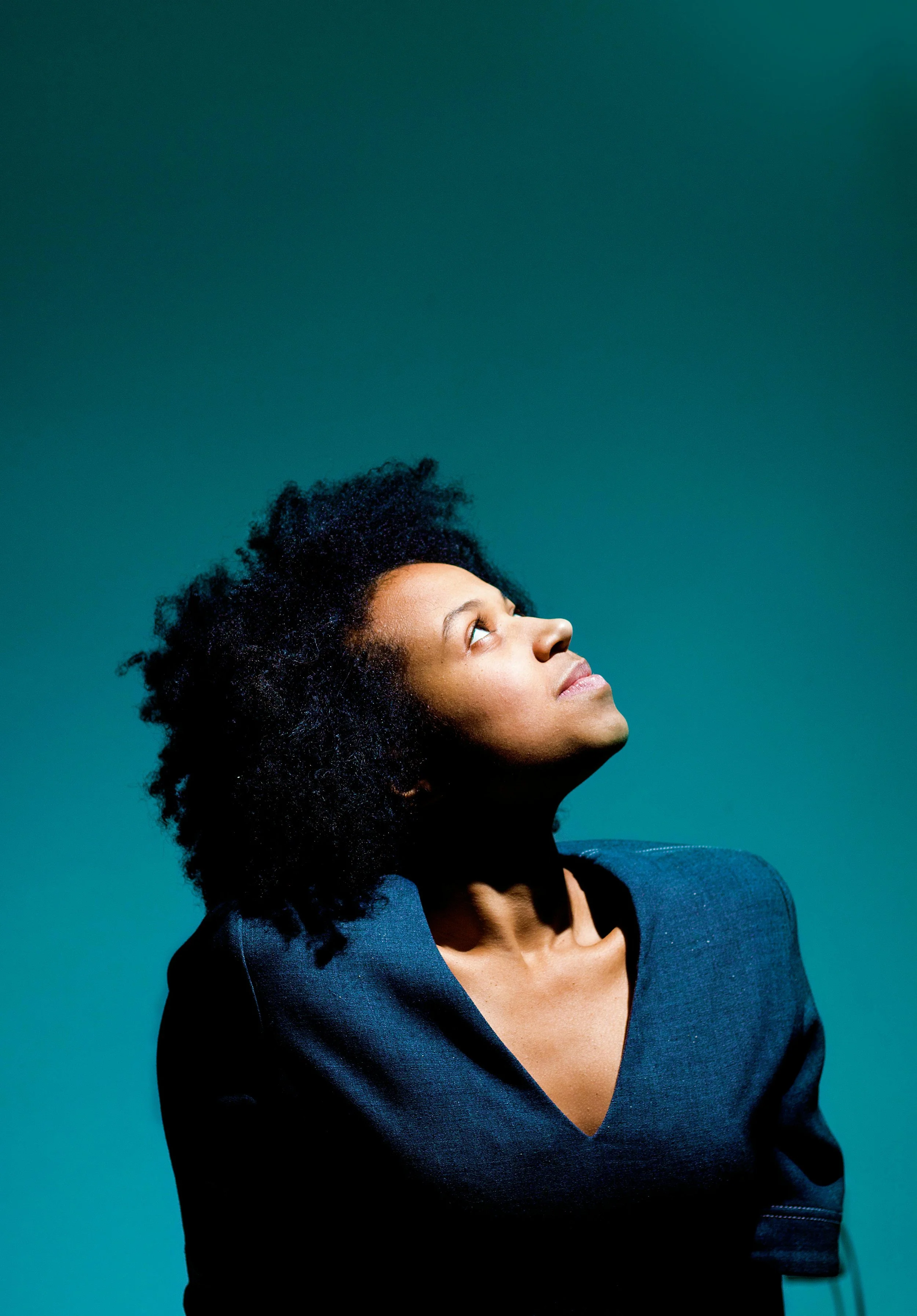 A woman with curly black hair looking upward against a teal background.