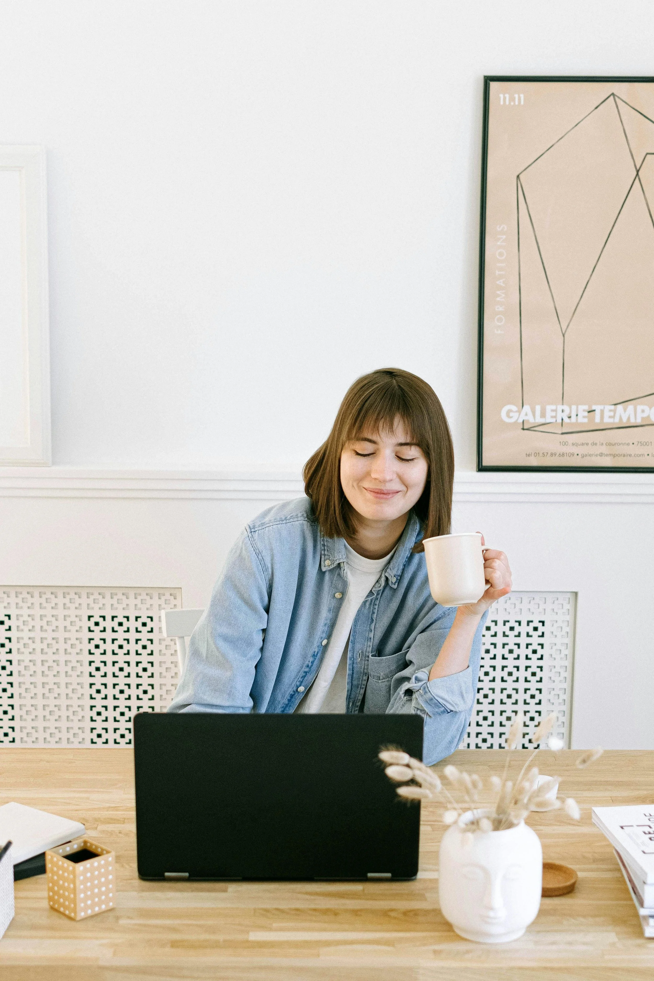 A young woman sitting at a wooden table with a closed black laptop in front of her. She is holding a white mug in her right hand, has shoulder-length brown hair, and is wearing a light denim shirt over a white t-shirt. She appears to be smiling with her eyes closed. On the table, there is a white vase with dried plants, a small dotted container, and some notebooks. The background features a white wall with a framed abstract geometric art piece and a decorative white panel.