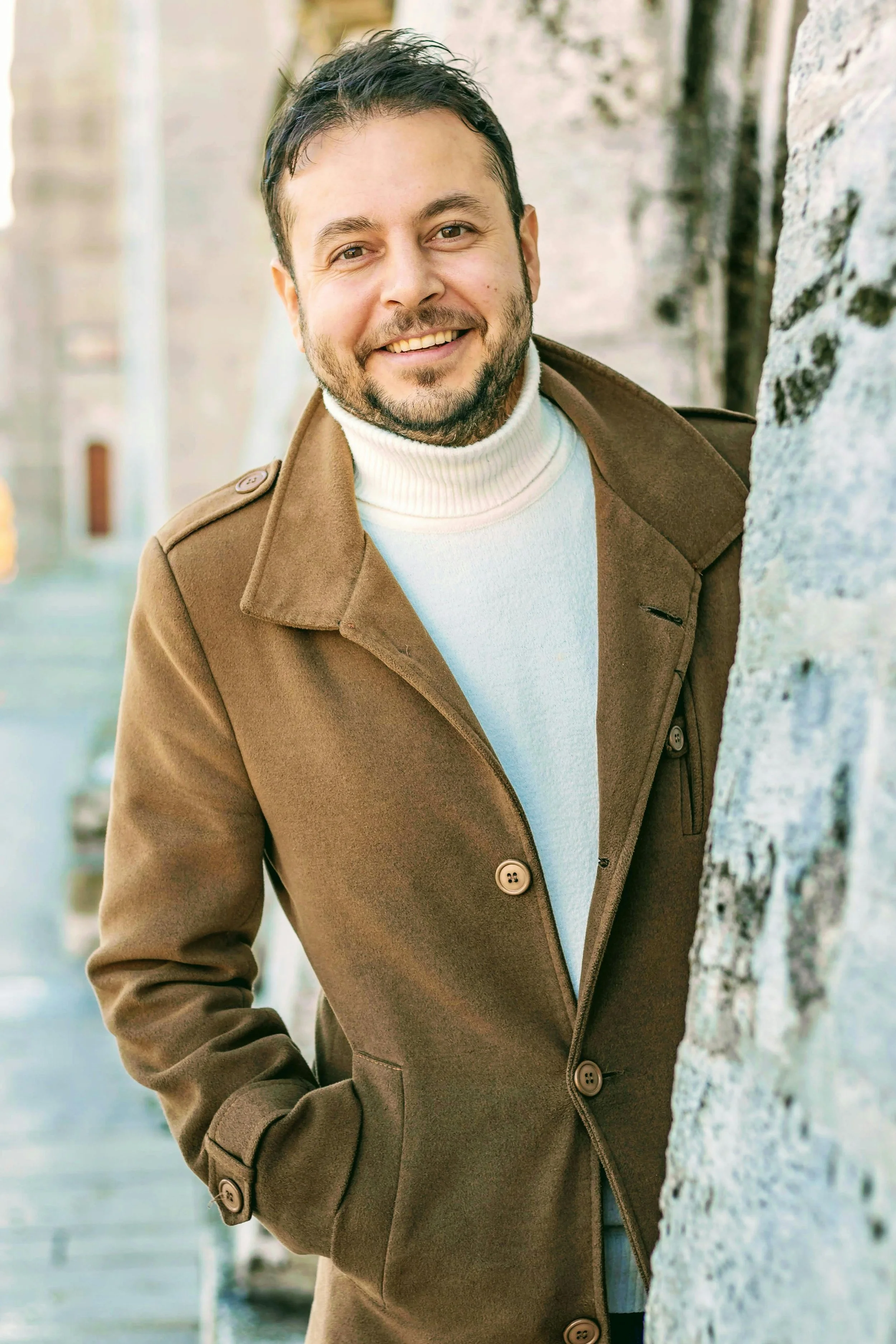 A man with dark hair and a beard smiling, wearing a brown coat and a white turtleneck, standing outdoors next to a stone wall.