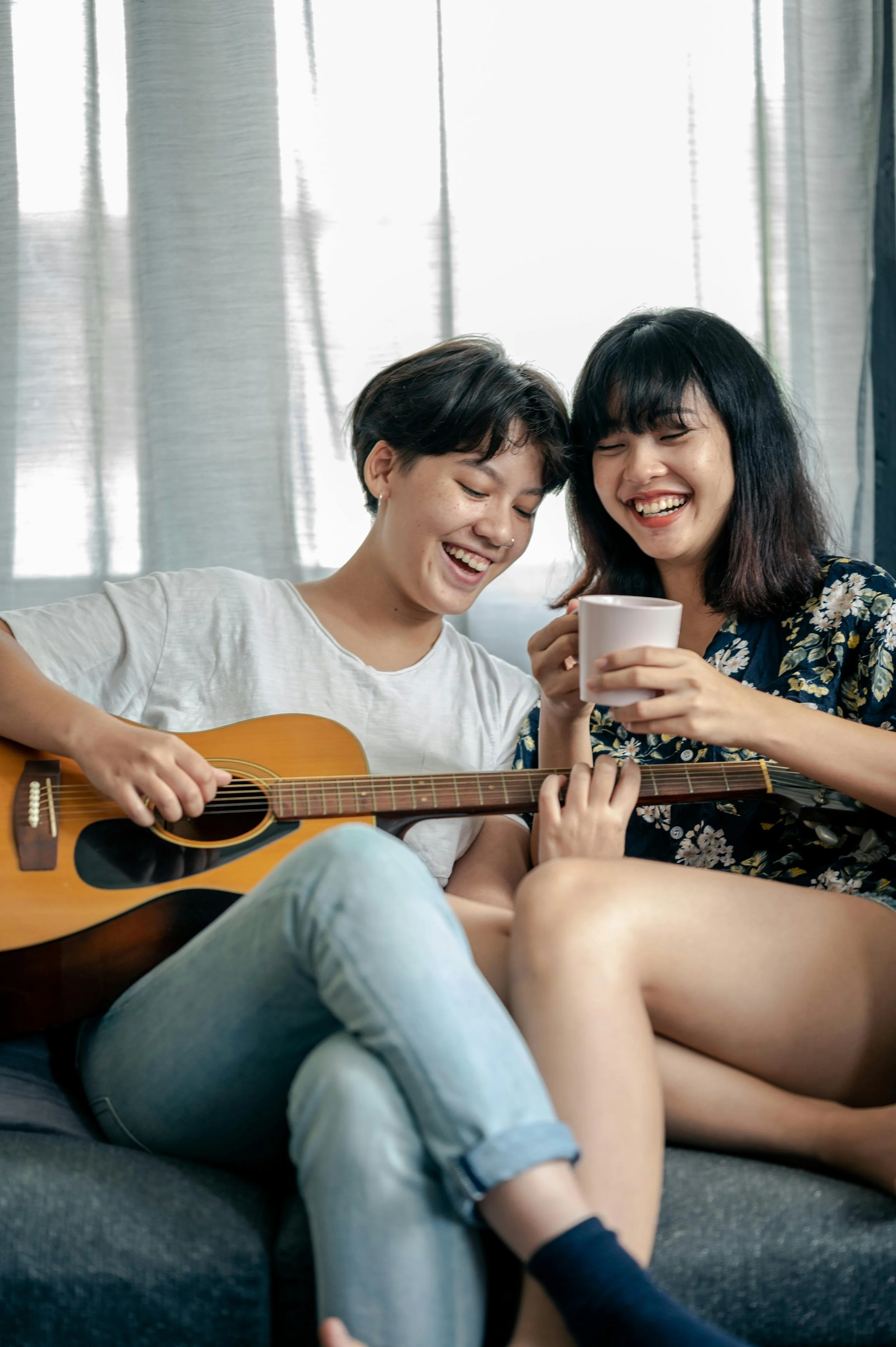 Two women sitting on a couch, one playing guitar and the other holding a mug, smiling and enjoying each other's company.