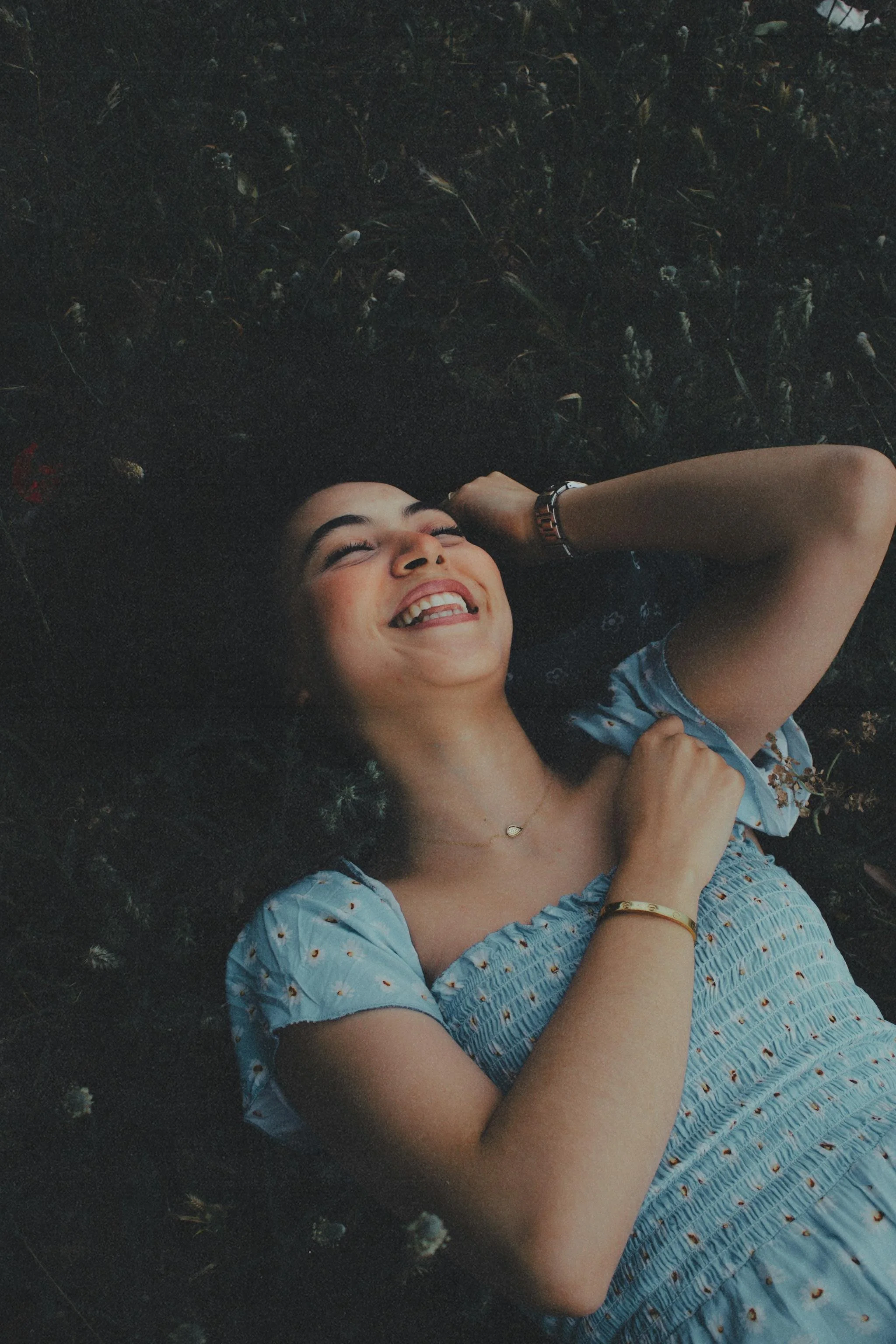 A young woman with dark hair lying on the ground, smiling and laughing, wearing a light blue dress with small patterns, a necklace, and gold bracelets.