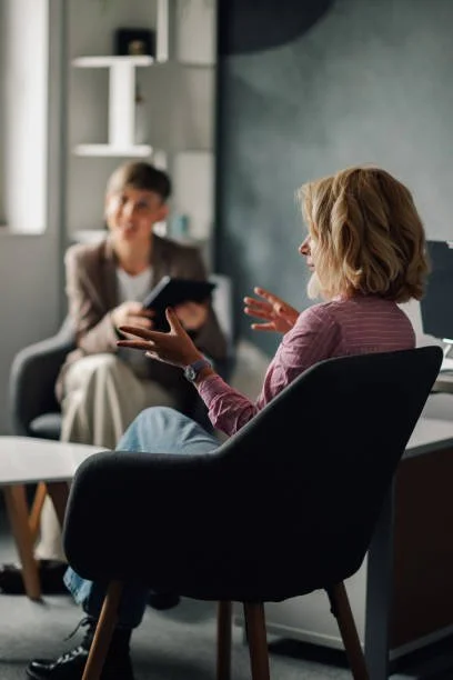 Two women having a conversation in a modern office or therapy room, one sitting in a black chair and the other in a couch, with bookshelf and window in the background.