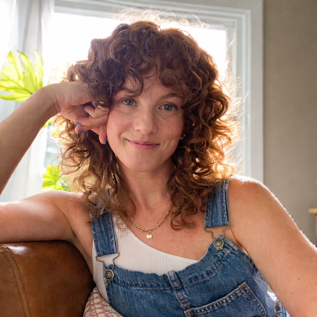 A woman with curly hair sitting on a couch beside a window, smiling.