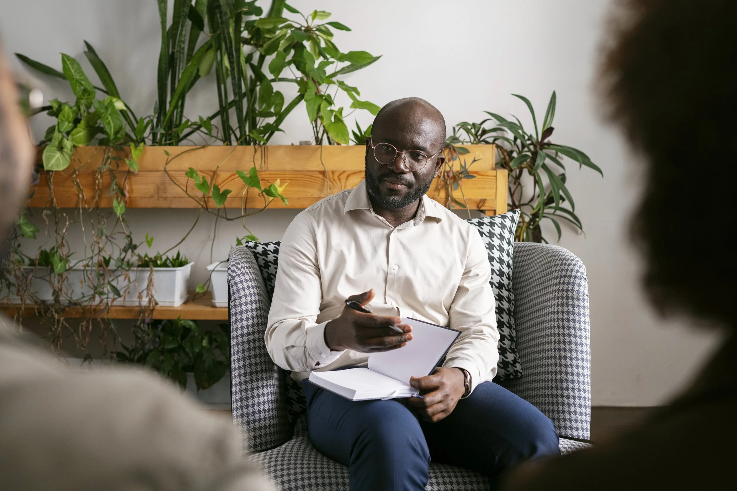 Man sitting in chair during a therapy or counseling session, holding a notebook and talking to two people in the foreground, with plants in the background.