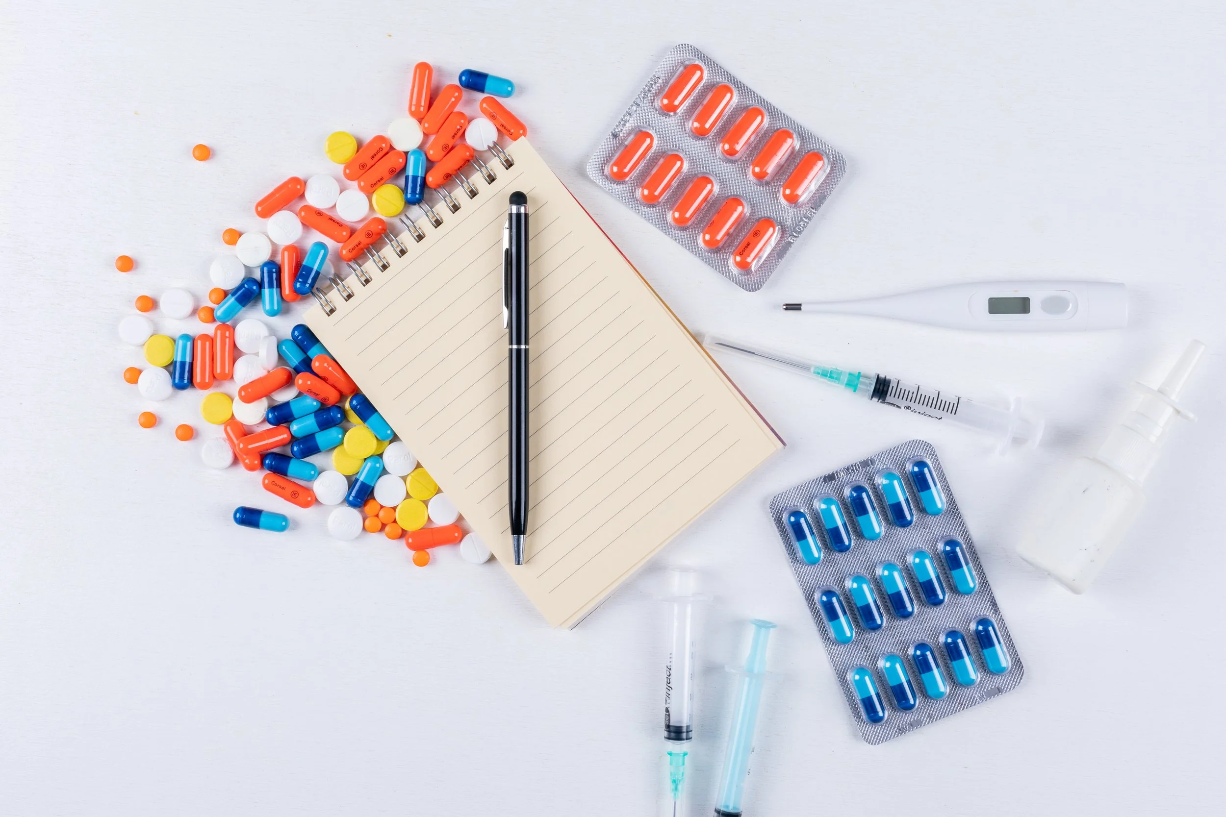 Various pills and capsules, a notebook with a pen, a digital thermometer, syringes, and medicine bottles on a white background, suggesting medication and healthcare tools.