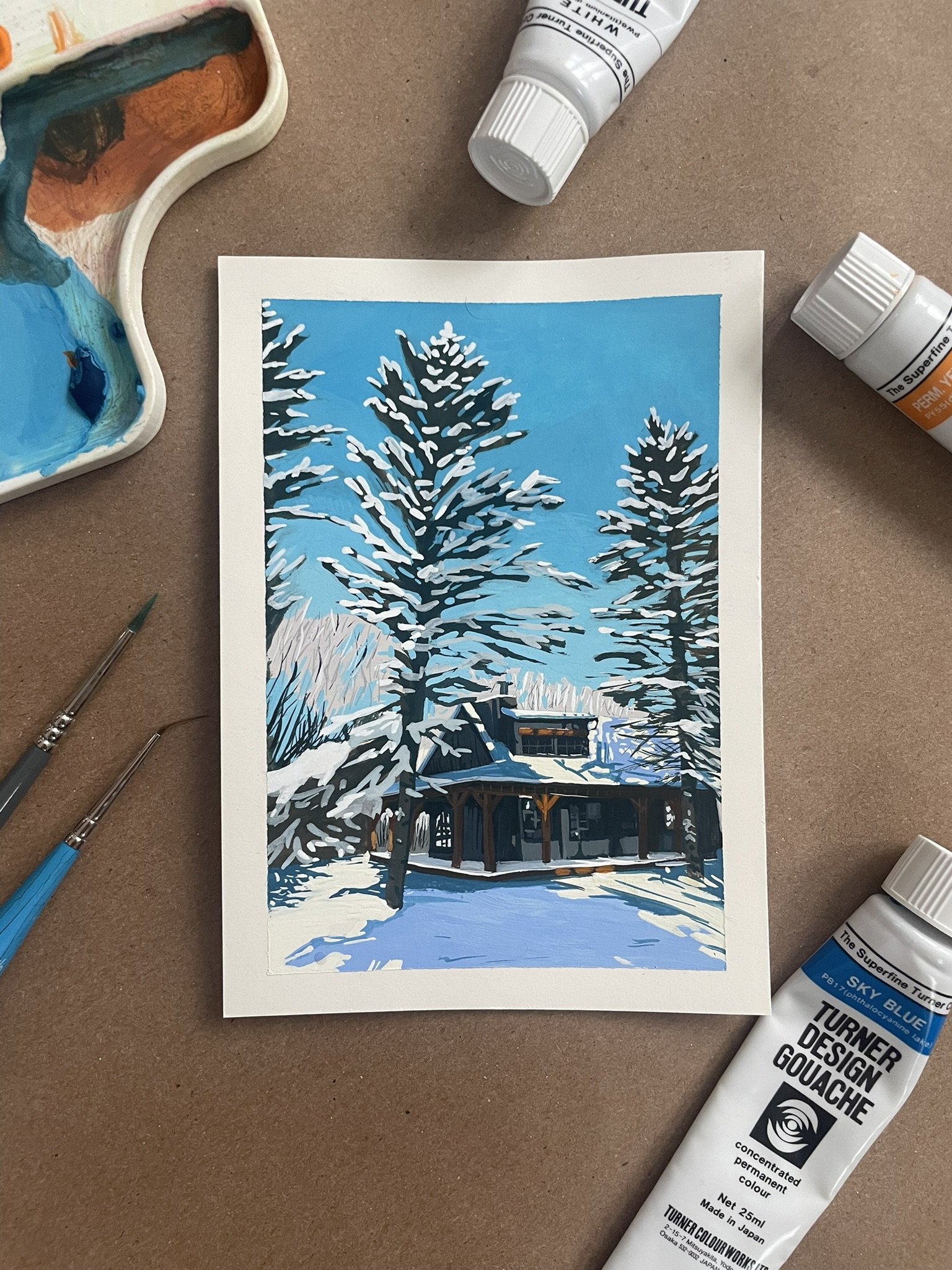 A painting of a snowy landscape in Riding Mountain National Park with tall pine trees and a cabin, created with gouache paints, surrounded by paint tubes and brushes.