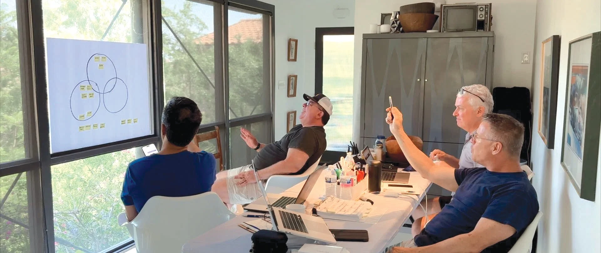 Four men seated at a long table in a bright room with large windows, engaged in a discussion or presentation. One person is taking a photo of a diagram on the window, while others are looking and listening. The table has laptops, notebooks, and office supplies, and there is a cabinet and artwork on the walls.