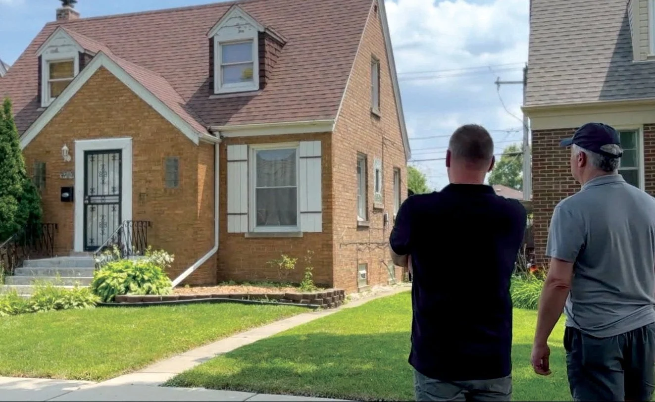 Two men standing on a sidewalk, facing a brick house with white shutters and a small set of stairs leading to the front door, in a residential neighborhood with a well-maintained lawn and cloudy sky.