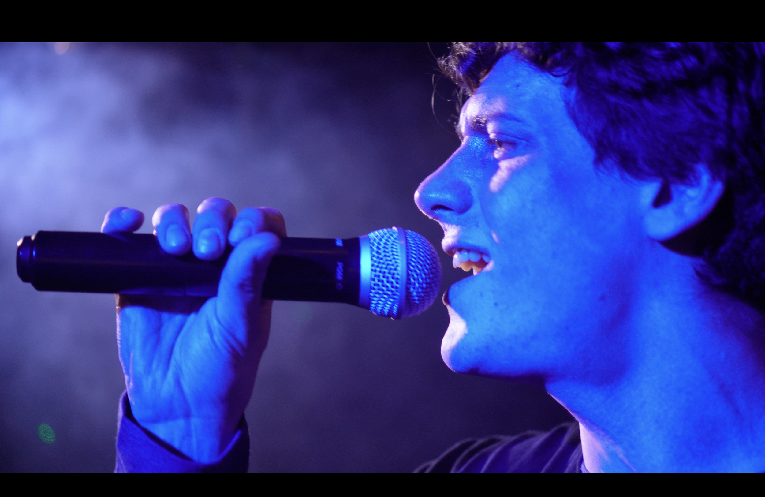 En person som sjunger in i en mikrofon på scen, med blå ljus belysning.