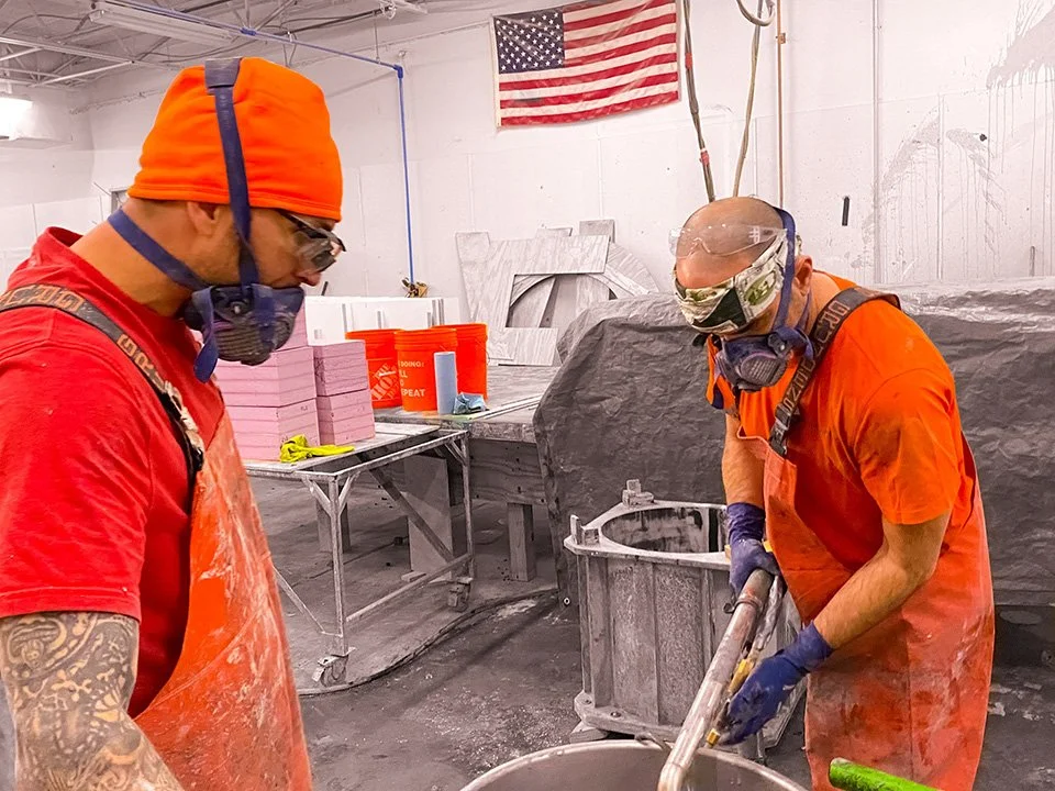 Two workers wearing orange shirts, orange head coverings, goggles, face masks, and gloves are working with a tool in a workshop. An American flag hangs on the wall behind them. The workspace has a metal table with pink foam blocks and an orange bucke