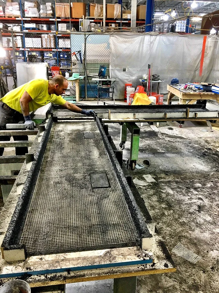 Worker in a yellow shirt and gloves working on a metal-framed screen or grate in an industrial warehouse or manufacturing facility.