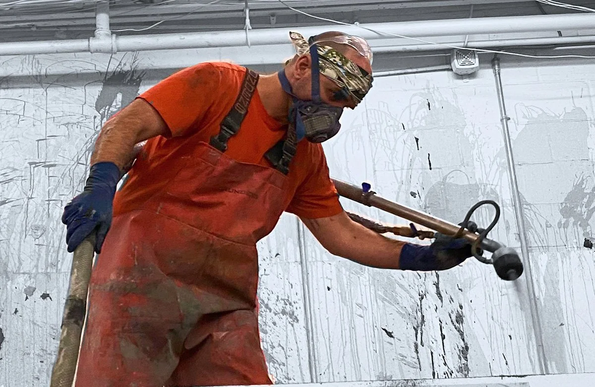 A man wearing protective gear and a mask is using a power tool to paint a wall.