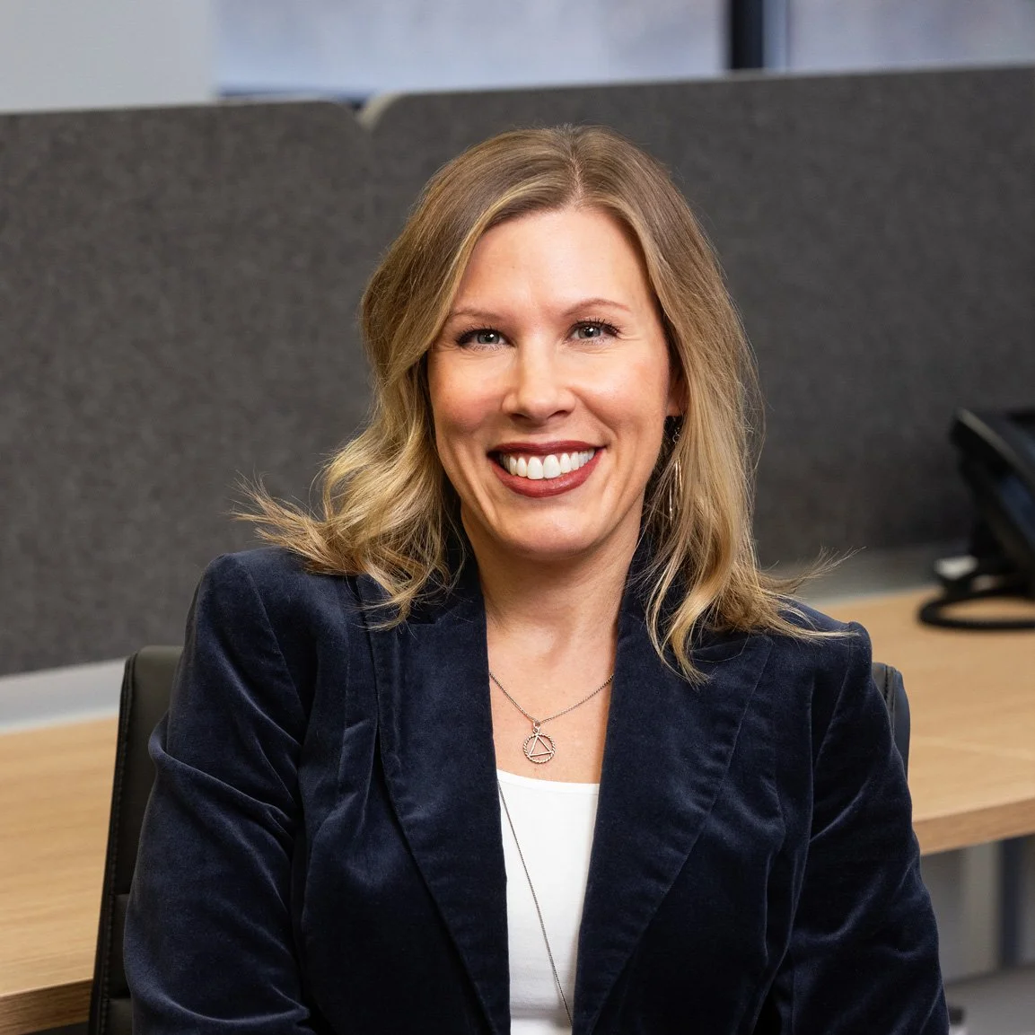 A woman with shoulder length blonde hair, wearing a blue jacket, sits at a desk in an office environment, smiling at the camera.