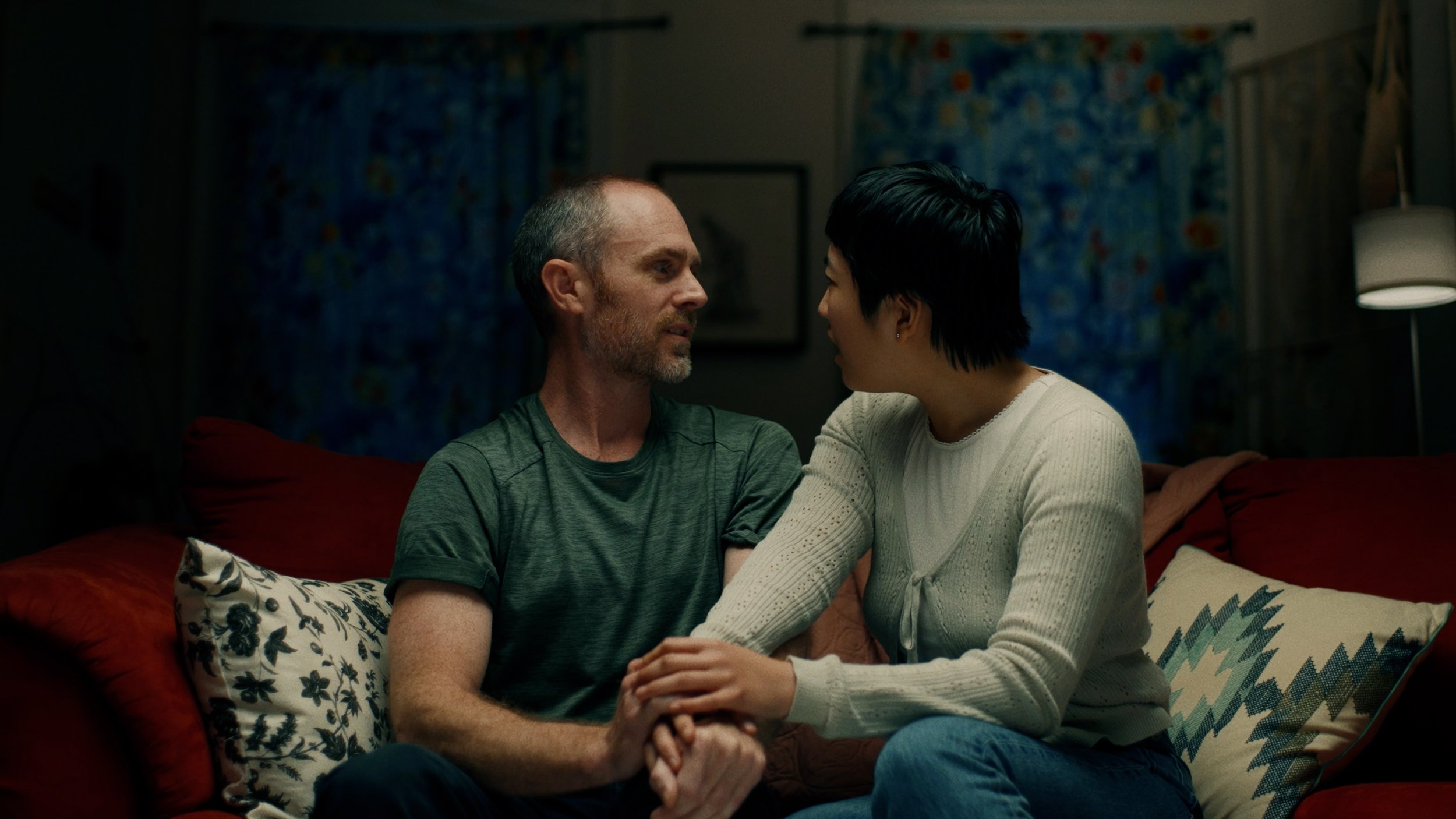 A couple sitting on a couch in a dimly lit room, engaged in conversation. The man is wearing a green T-shirt and the woman is wearing a white sweater. They are sitting close together with their hands gently touching. The couch has decorative patterne