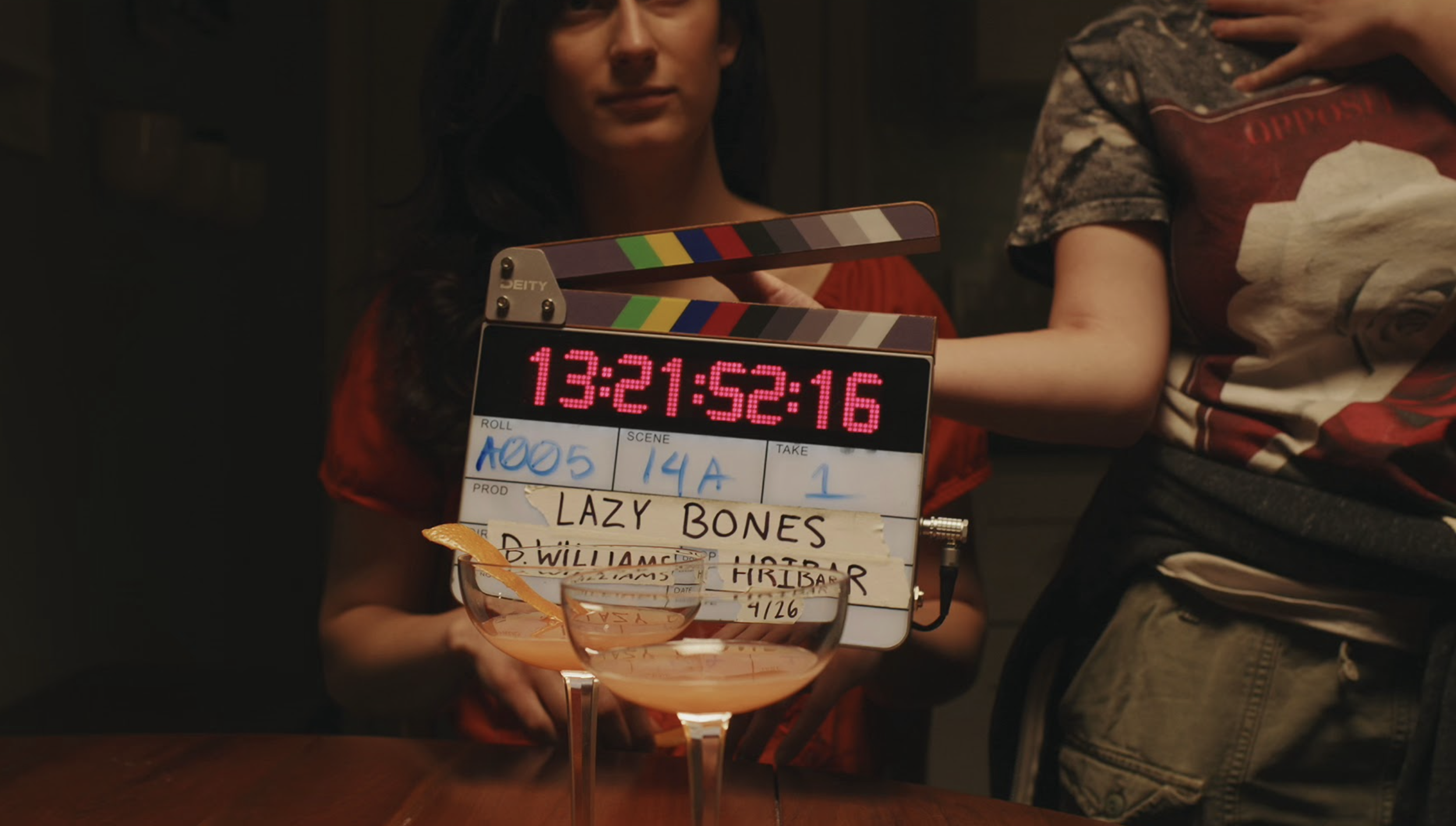 Film clapperboard labeled 'Lazy Bones' with two people partially visible, one in a red shirt and the other wearing a graphic t-shirt and jeans.