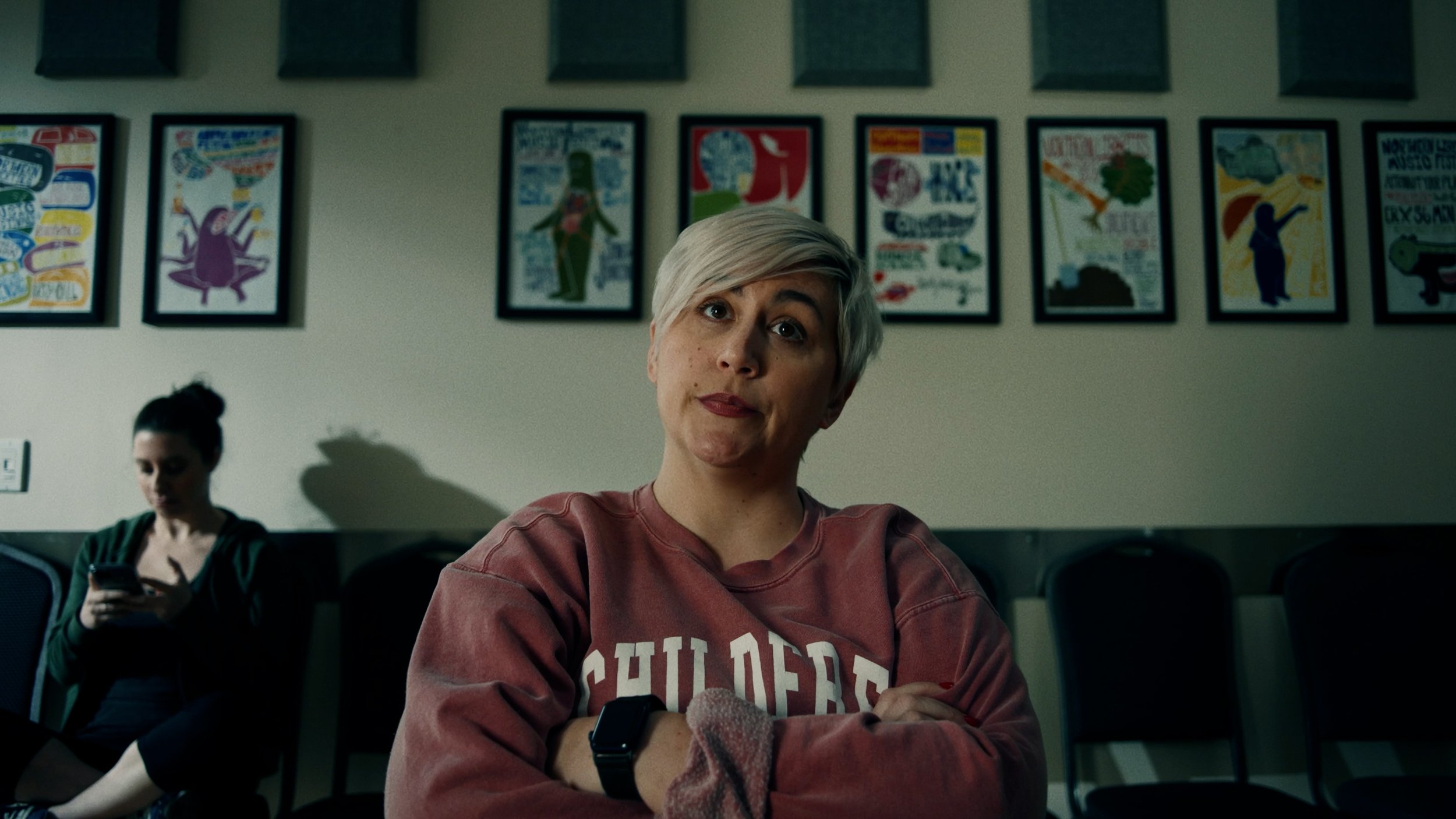 Woman sitting in a room with posters on the wall, wearing a sweatshirt with "CHILDREN" text.