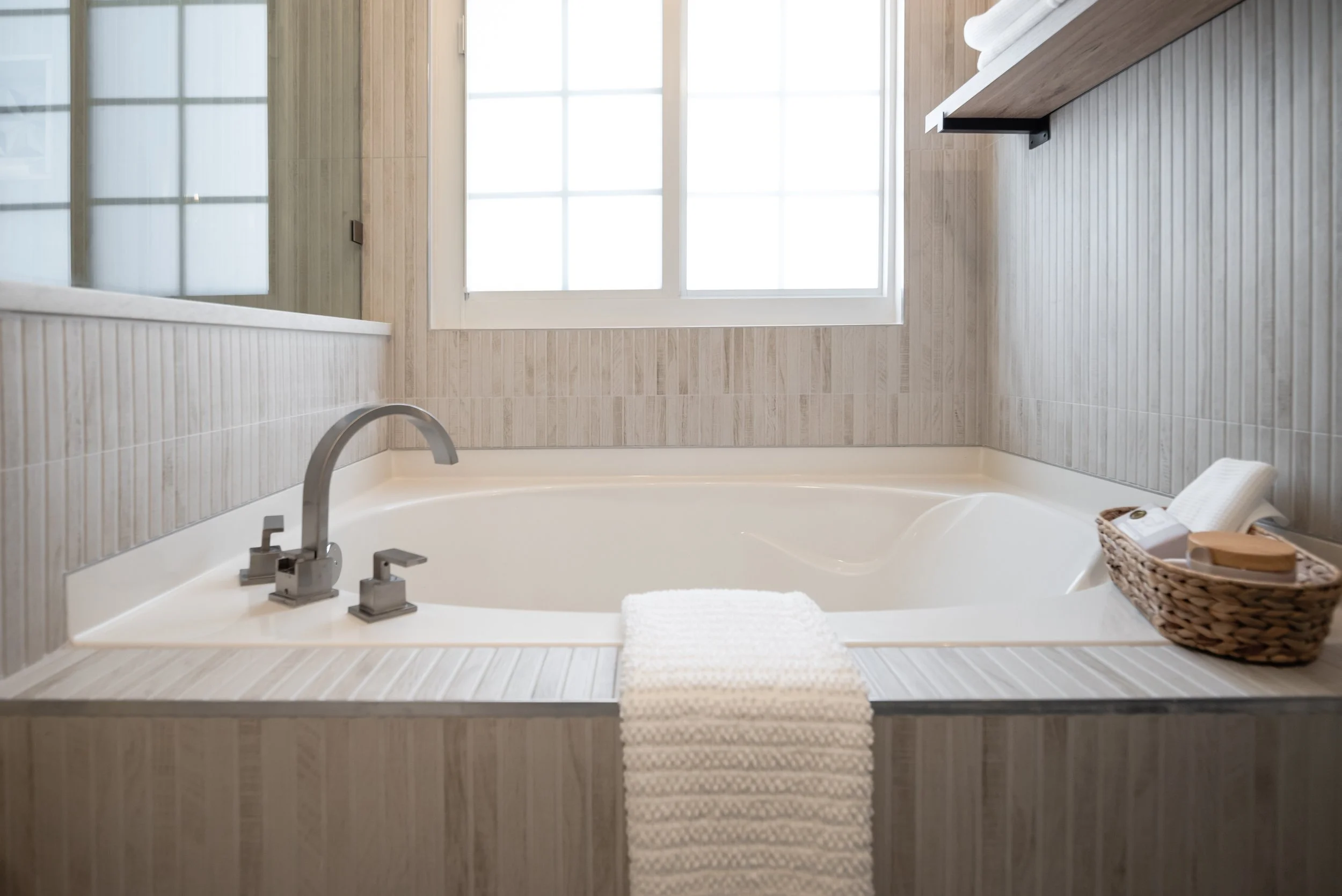 A modern bathroom with a built-in bathtub near a large window, tiled walls, a white towel on the edge, and a wicker basket with toiletries.