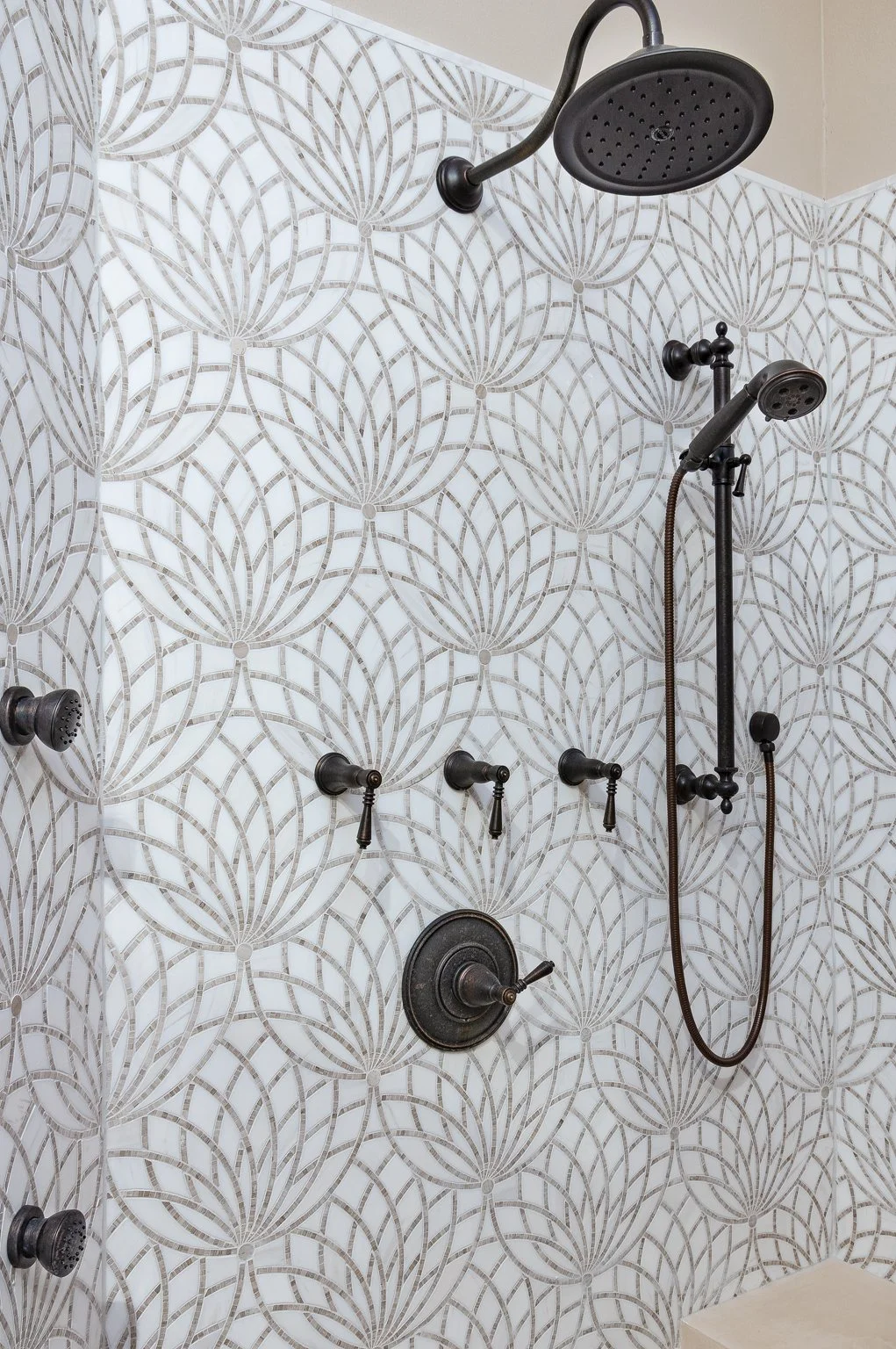 A shower with patterned tiles featuring a repetitive floral motif in neutral tones. The shower has multiple fixtures, including a large overhead rain shower, a handheld showerhead, and three wall-mounted knobs for controlling water flow and temperature.