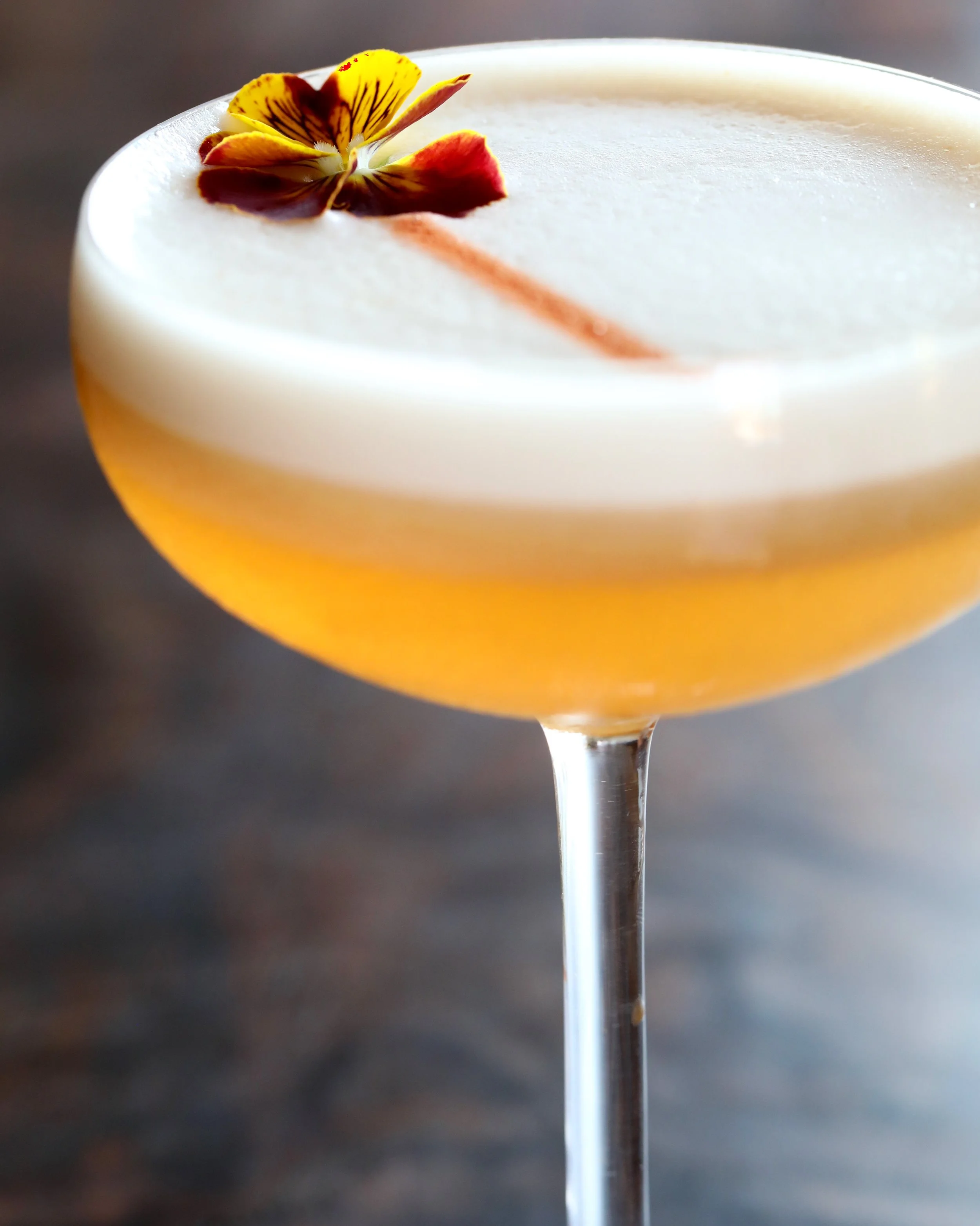 A cocktail drink topped with a yellow and red edible flower, set against a dark background.
