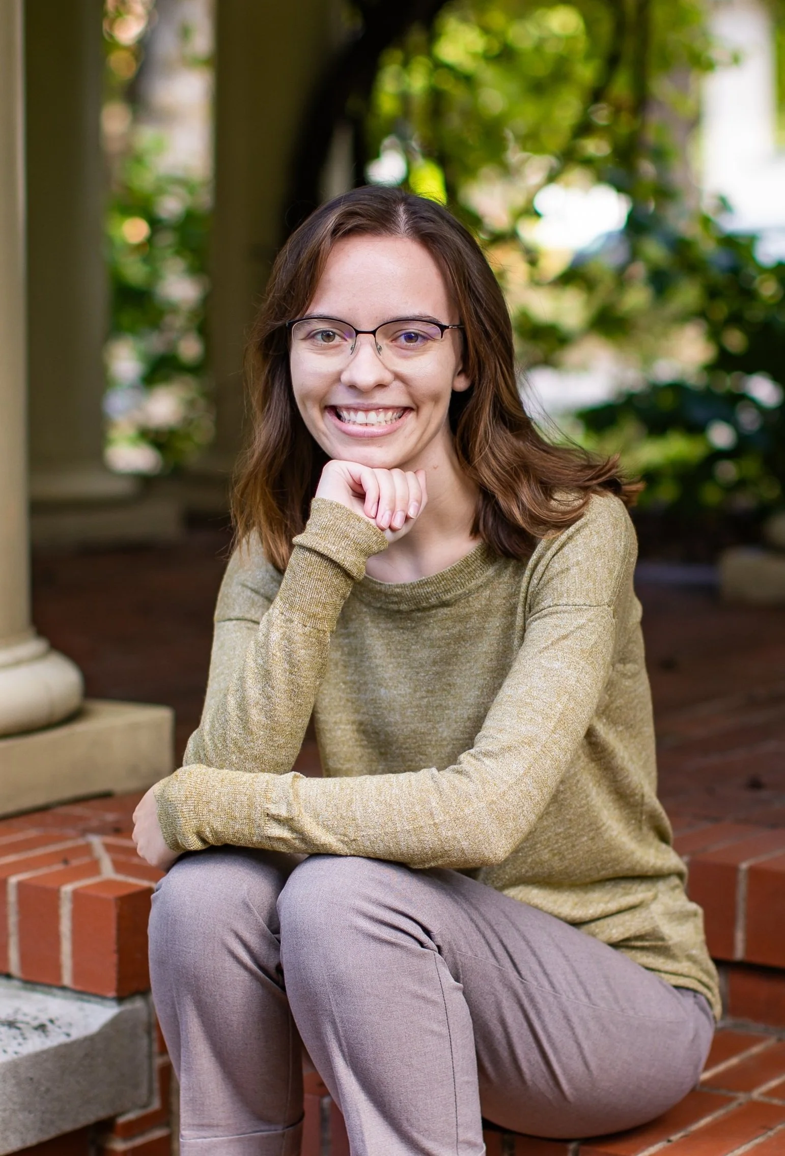 A young woman with glasses, shoulder-length brown hair, wearing a yellow-green sweater and light pink pants, sitting on a brick step outdoors, smiling at the camera.