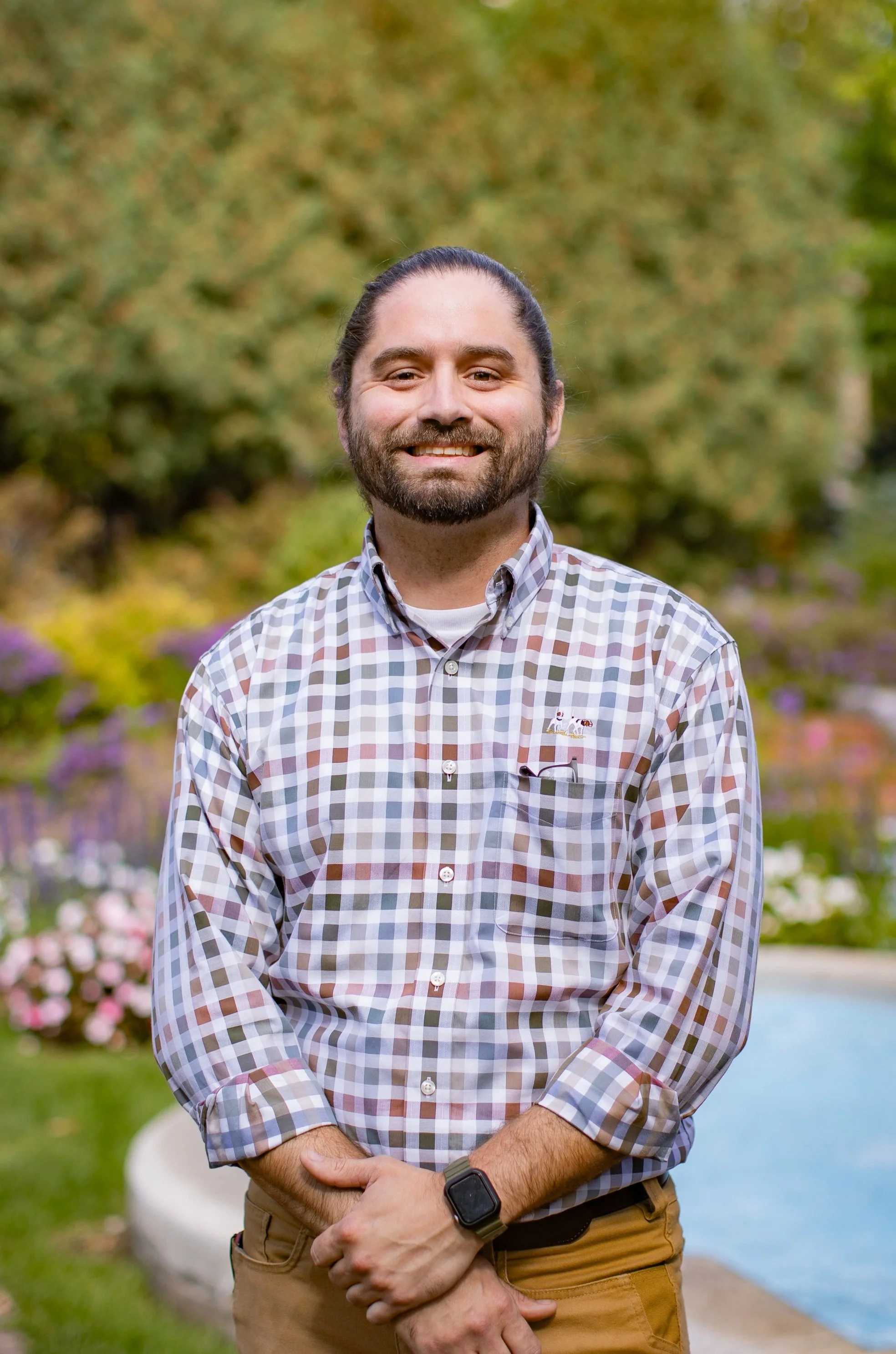 A man with long hair tied back, a beard, and a plaid shirt standing outdoors in a garden with colorful flowers and trees.