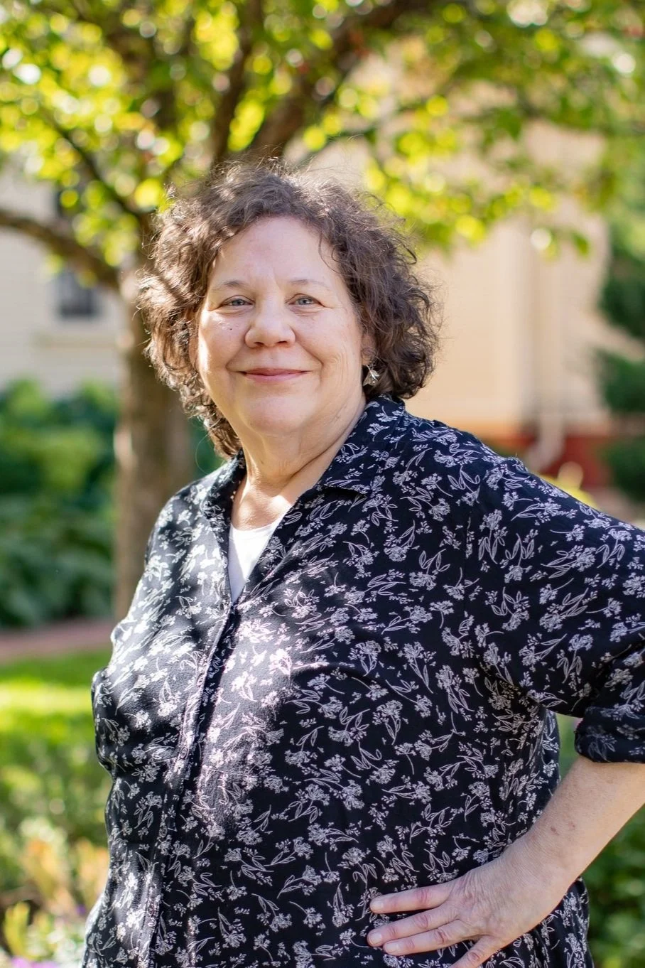 A smiling woman with short curly hair wearing a black and white floral shirt, standing outdoors in front of a tree with green leaves and a house in the background.