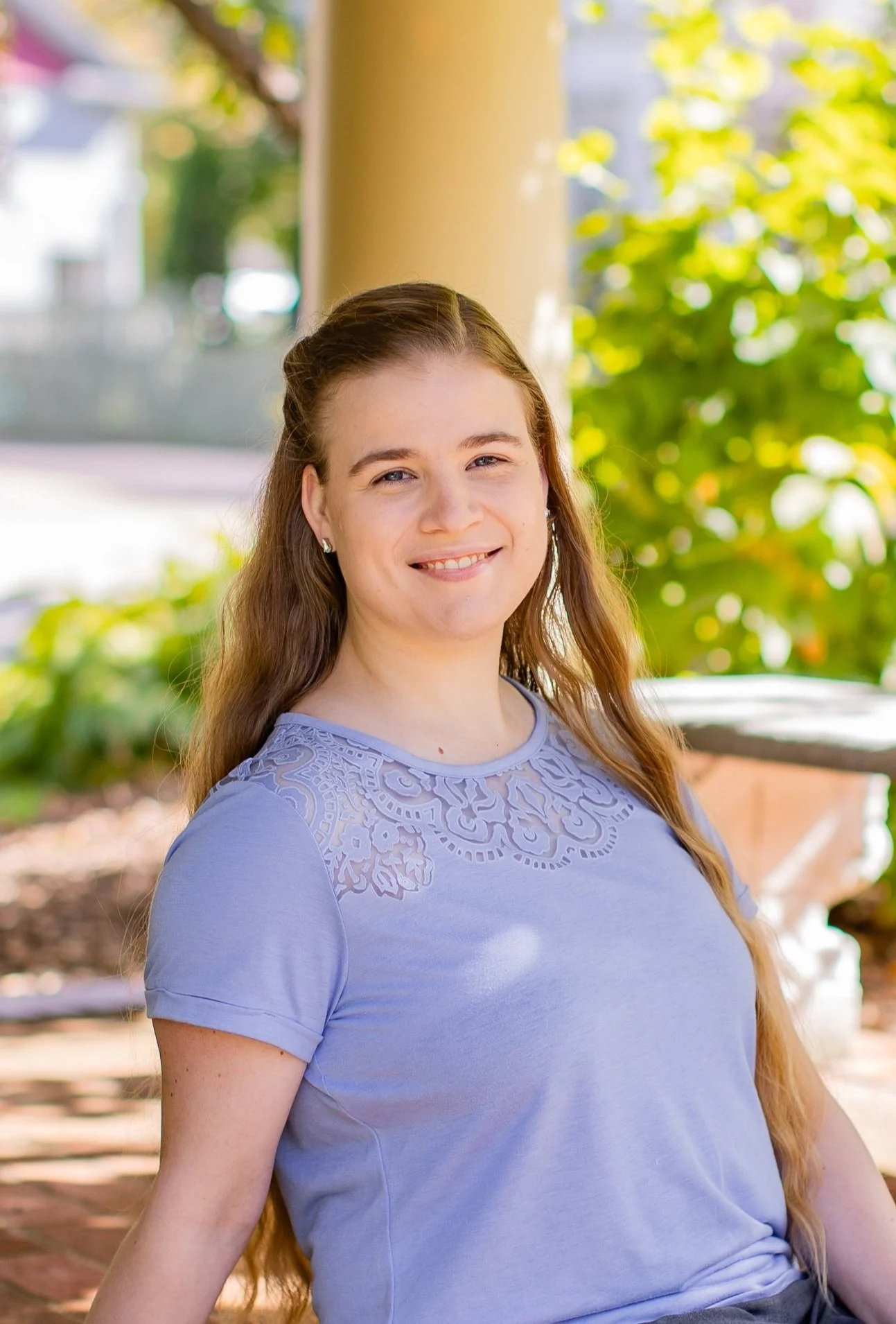 A young woman with long light brown hair and fair skin, wearing a lavender shirt with lace detailing, smiling outdoors on a sunny day with green foliage in the background.